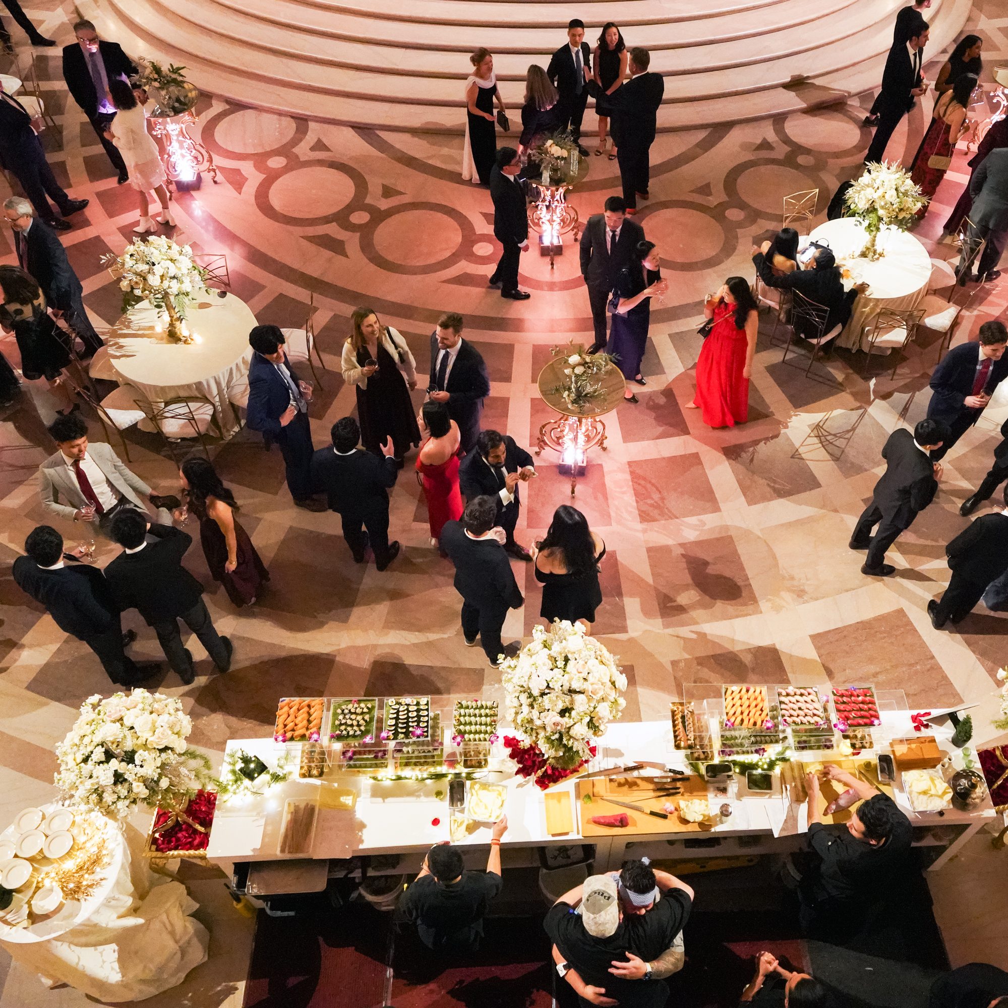 A formal gathering with elegantly dressed people mingling in a grand hall. Flowers and lavish food tables are arranged, creating a sophisticated atmosphere.