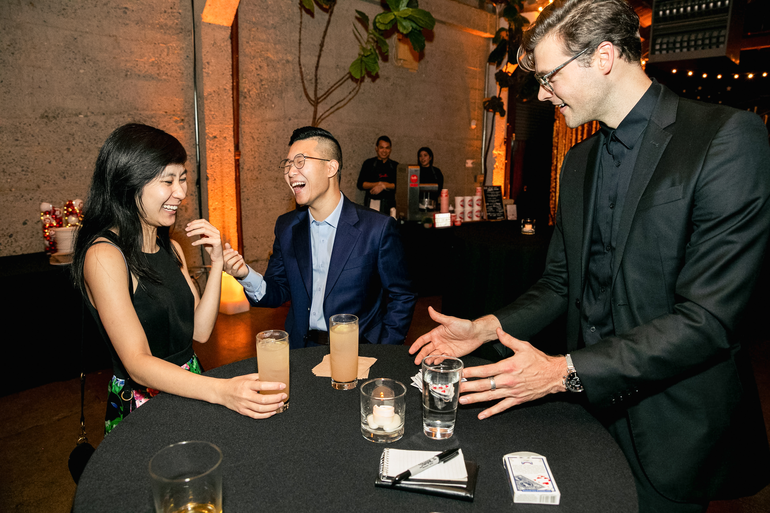 Three people gather around a table at a party, laughing. One man performs a magic trick with a card in a glass. Drinks and a notepad are on the table.