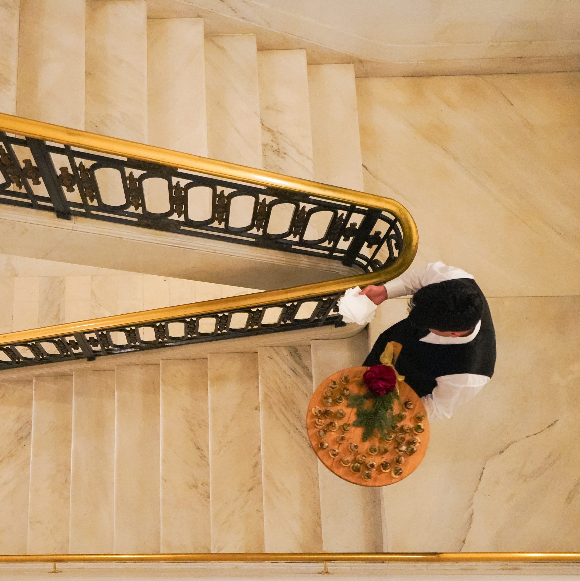 A person in a white shirt and black vest carries a tray with glasses and a rose down a marble staircase, bordered by an ornate gold and black railing.