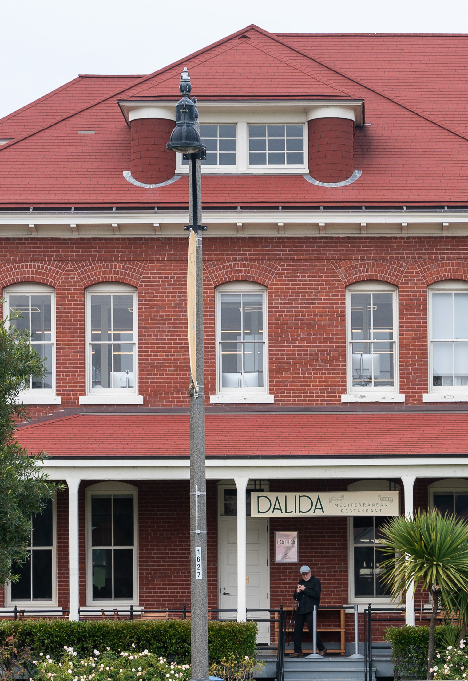 A person stands outside a red-brick building with a sign reading &quot;DALIDA Mediterranean Restaurant.&quot; There's a tall streetlamp and greenery nearby.