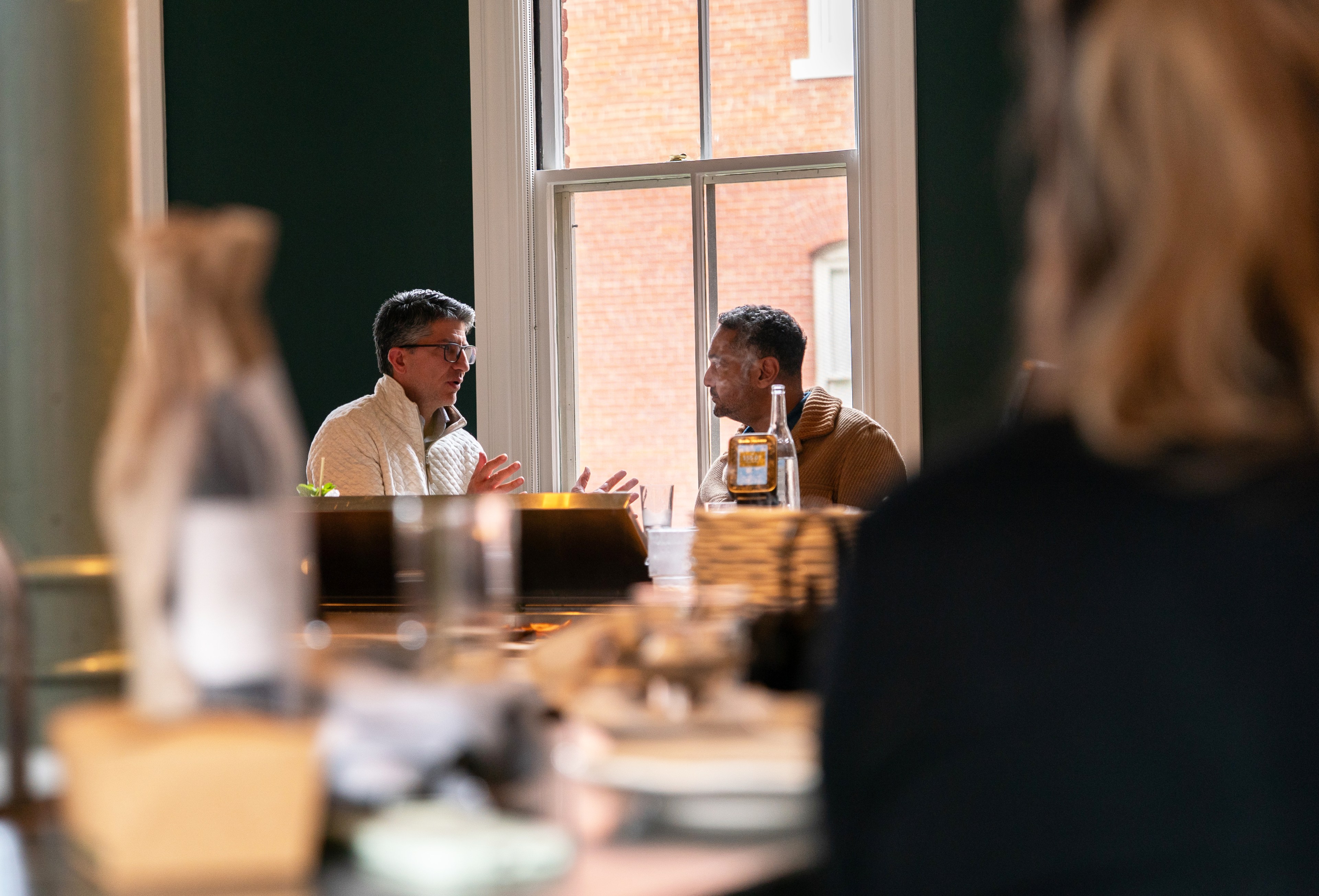 Two people sit at a table near a window, engaged in conversation. A clear bottle is on the table, with soft lighting in the background.