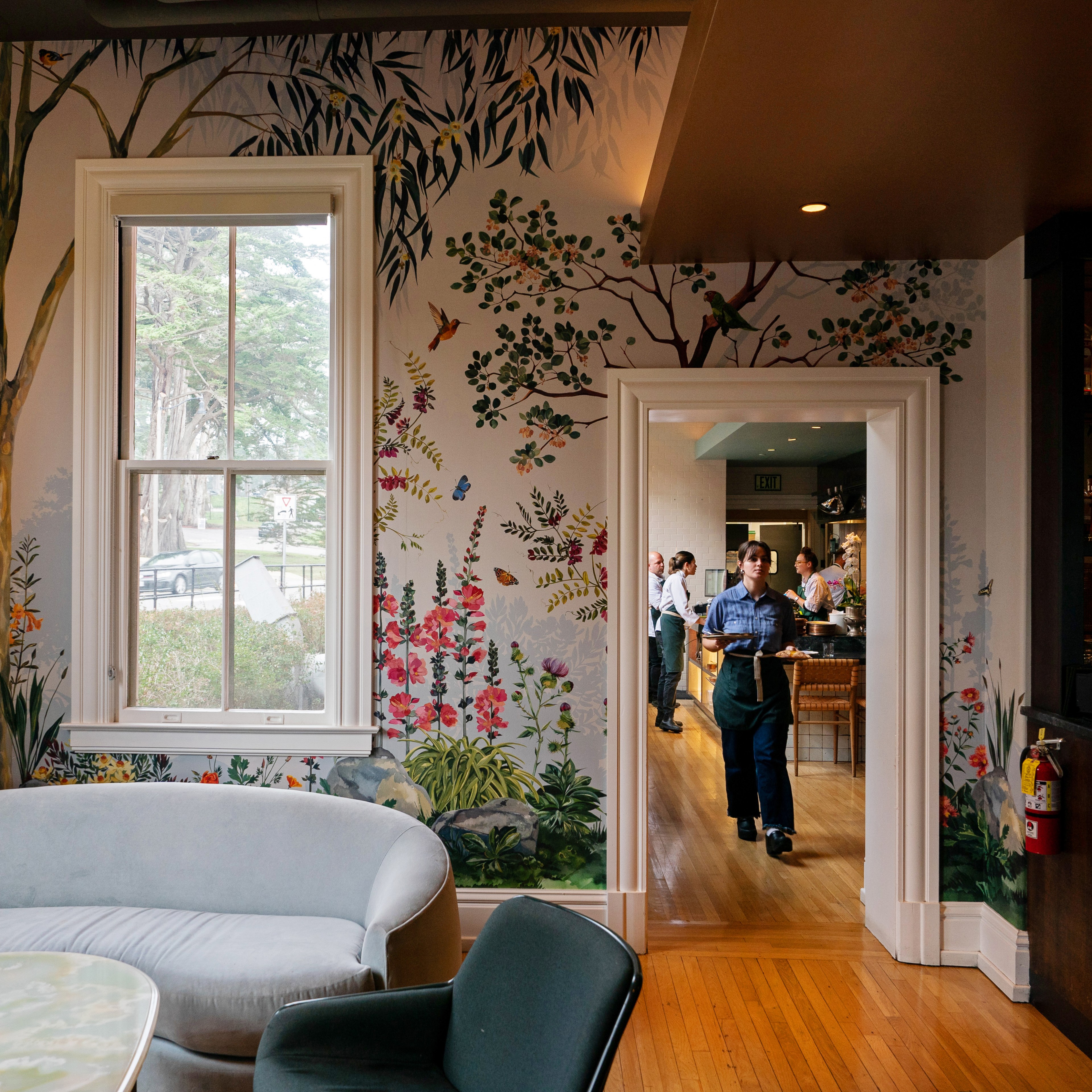 A cozy room with floral and bird murals, featuring a gray sofa and a person carrying drinks from an adjacent room. A bar with bottles is to the right.