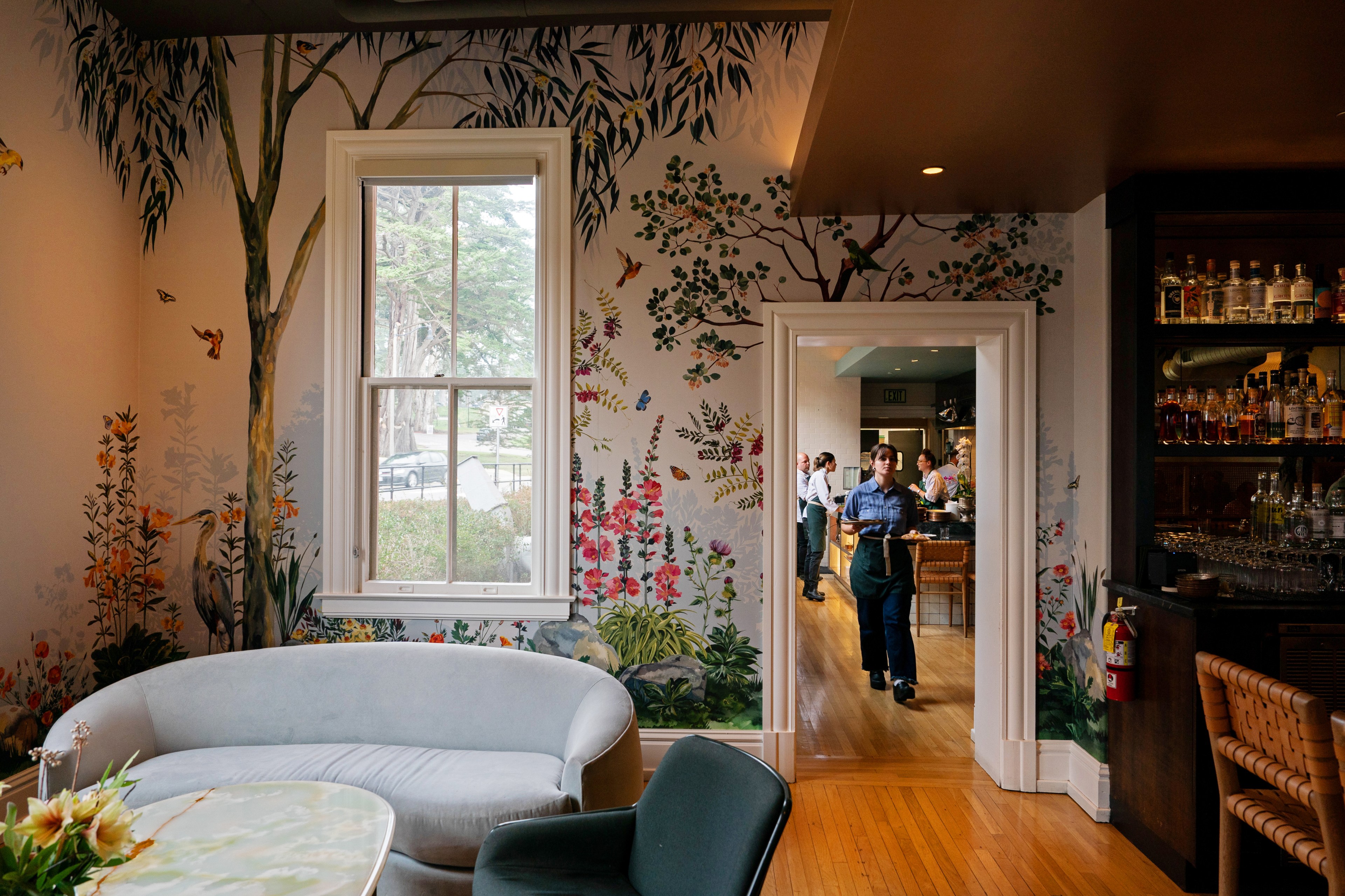 A cozy room with floral and bird murals, featuring a gray sofa and a person carrying drinks from an adjacent room. A bar with bottles is to the right.