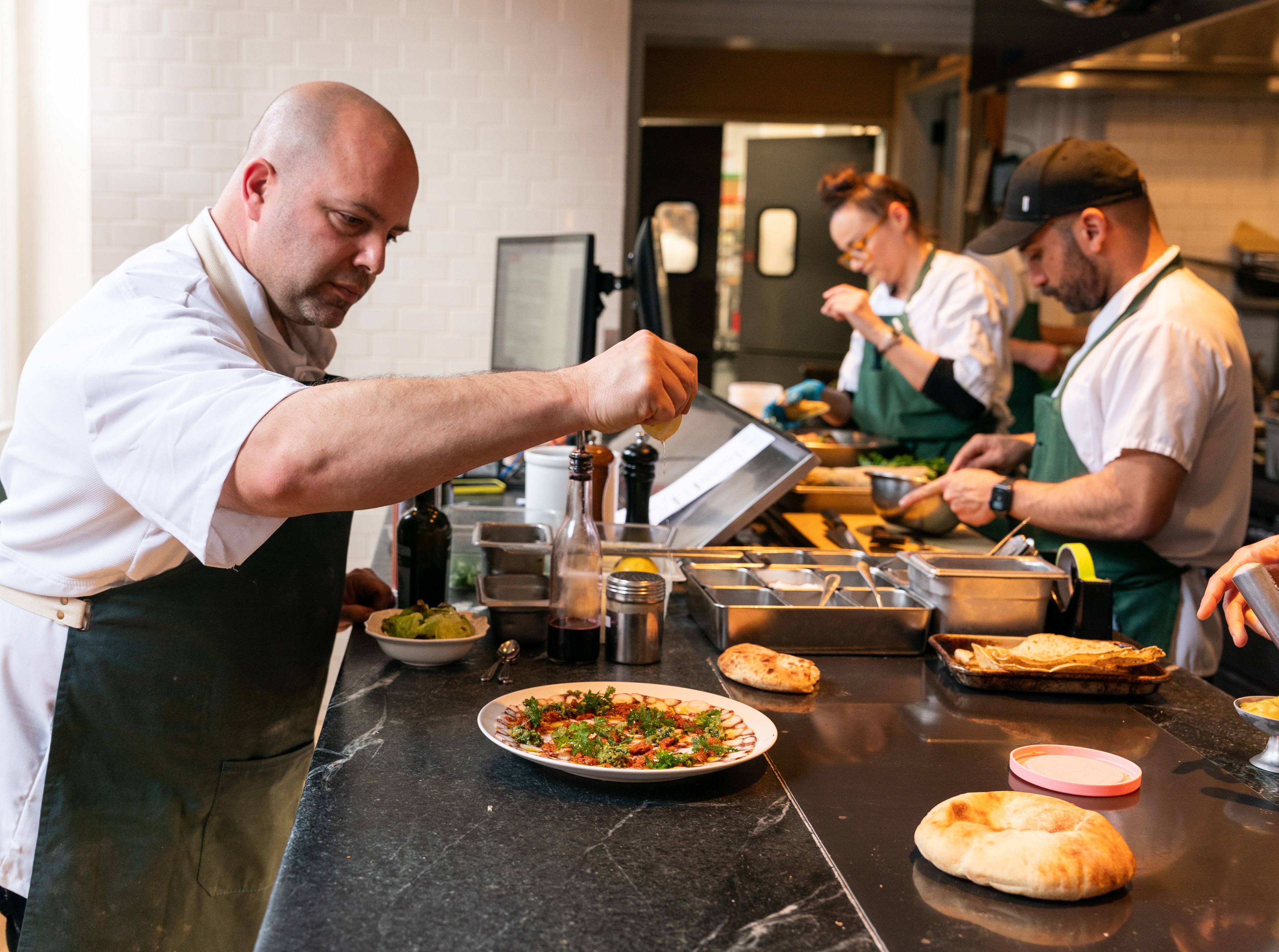 A chef sprinkles seasoning on a dish, surrounded by ingredients and utensils on a countertop. Two others in the background prepare food in a professional kitchen.