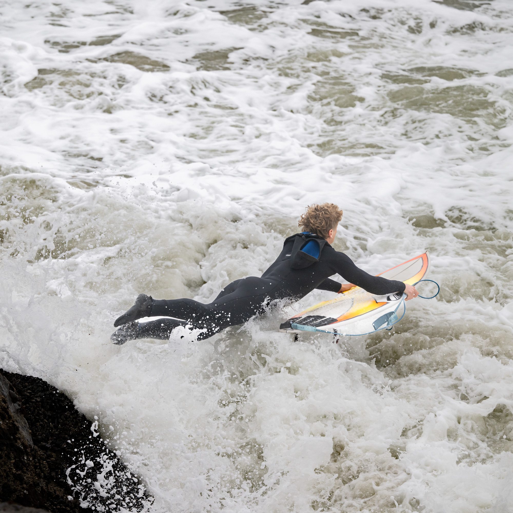 A surfer in a wetsuit is paddling on a surfboard through churning, foamy ocean waves, heading away from a rocky shoreline.