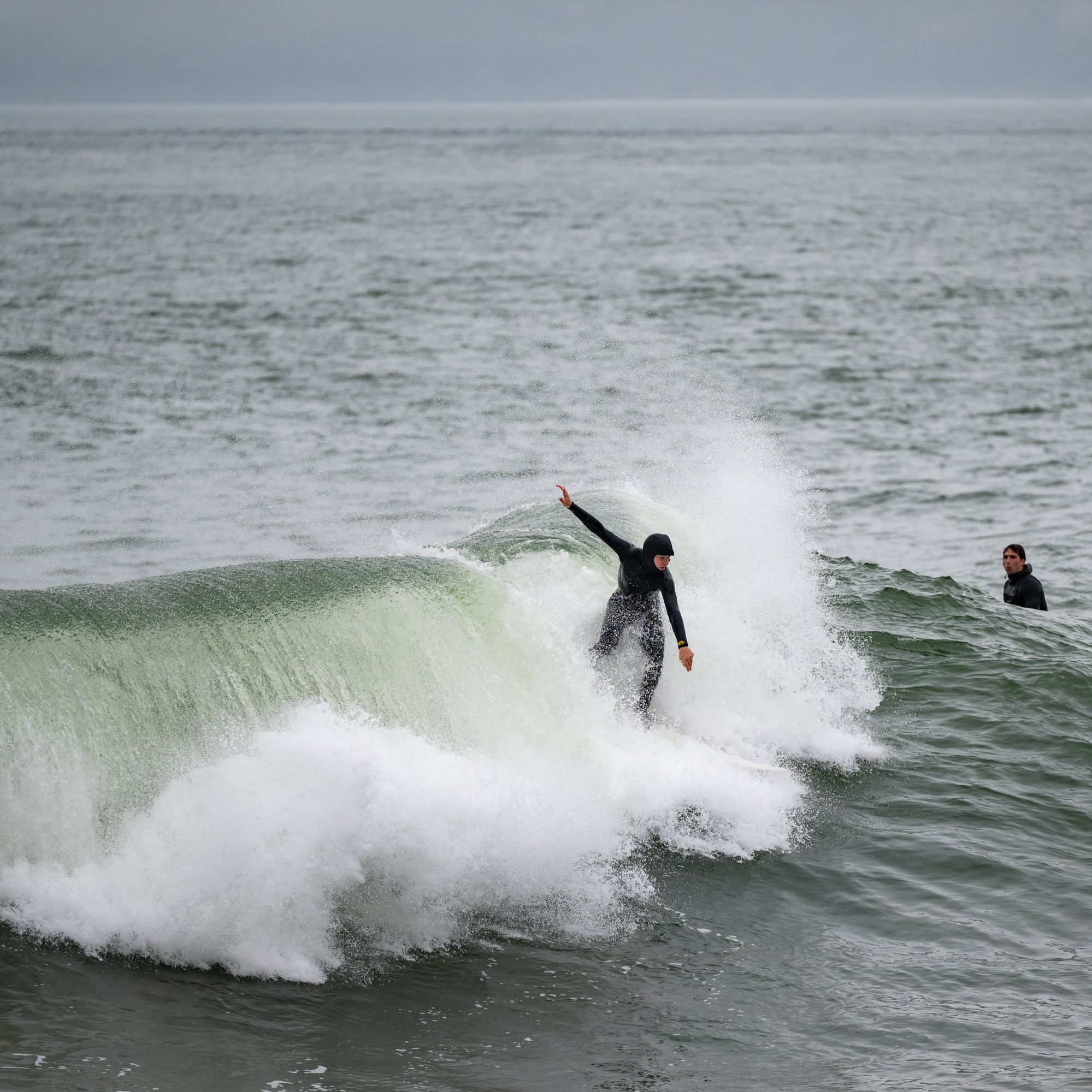 A surfer in a black wetsuit rides a breaking wave, skillfully balancing on a surfboard, while another person watches from the water's surface nearby.