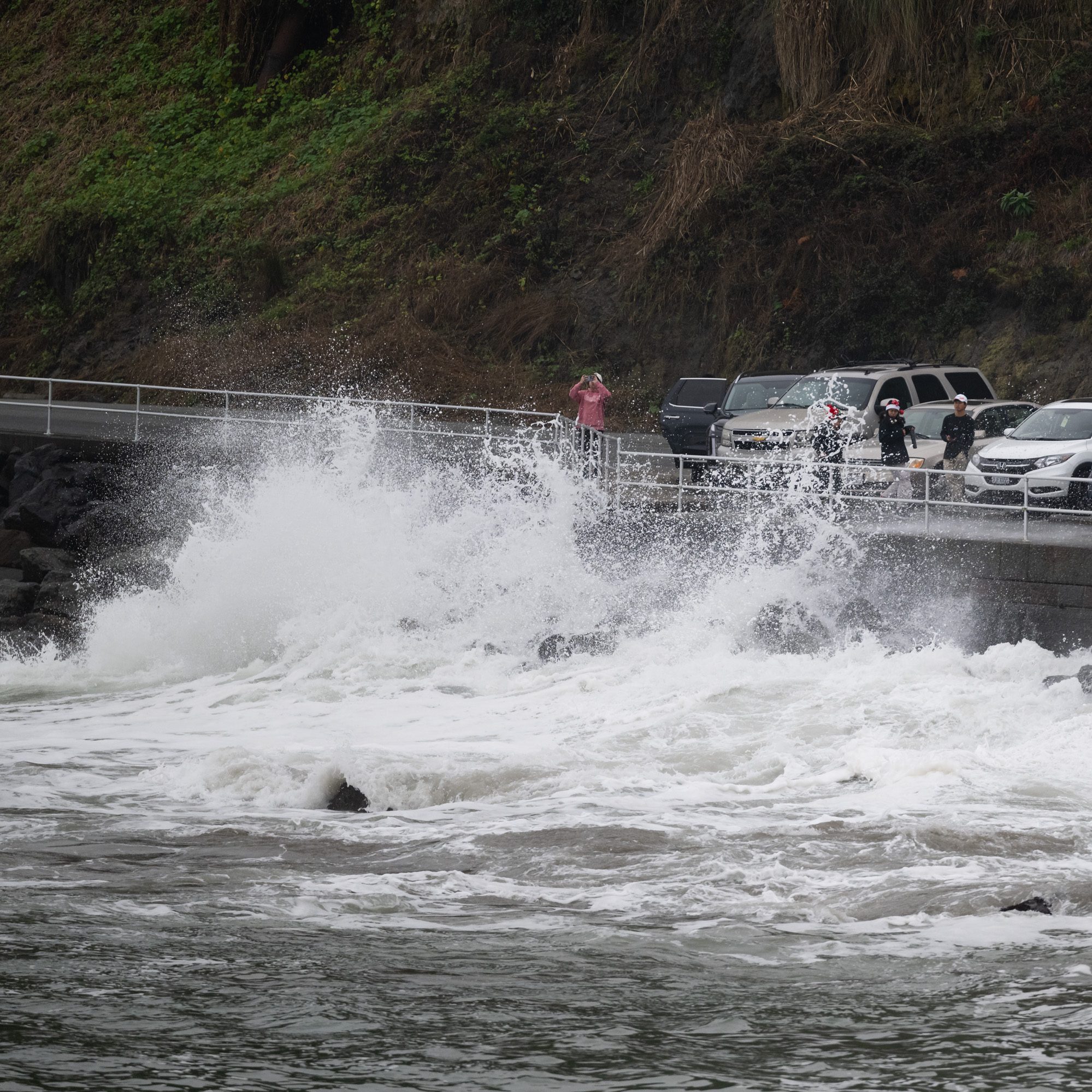 Waves crash forcefully against a seawall, splashing water high. Nearby, people stand on a walkway taking photos, with parked cars and green cliffs in the background.