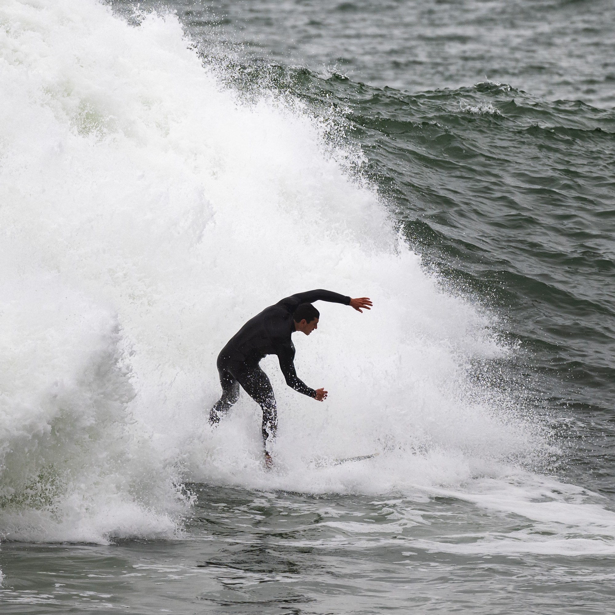 A surfer in a black wetsuit rides a powerful, frothy wave in the ocean, with water splashing dramatically around them.