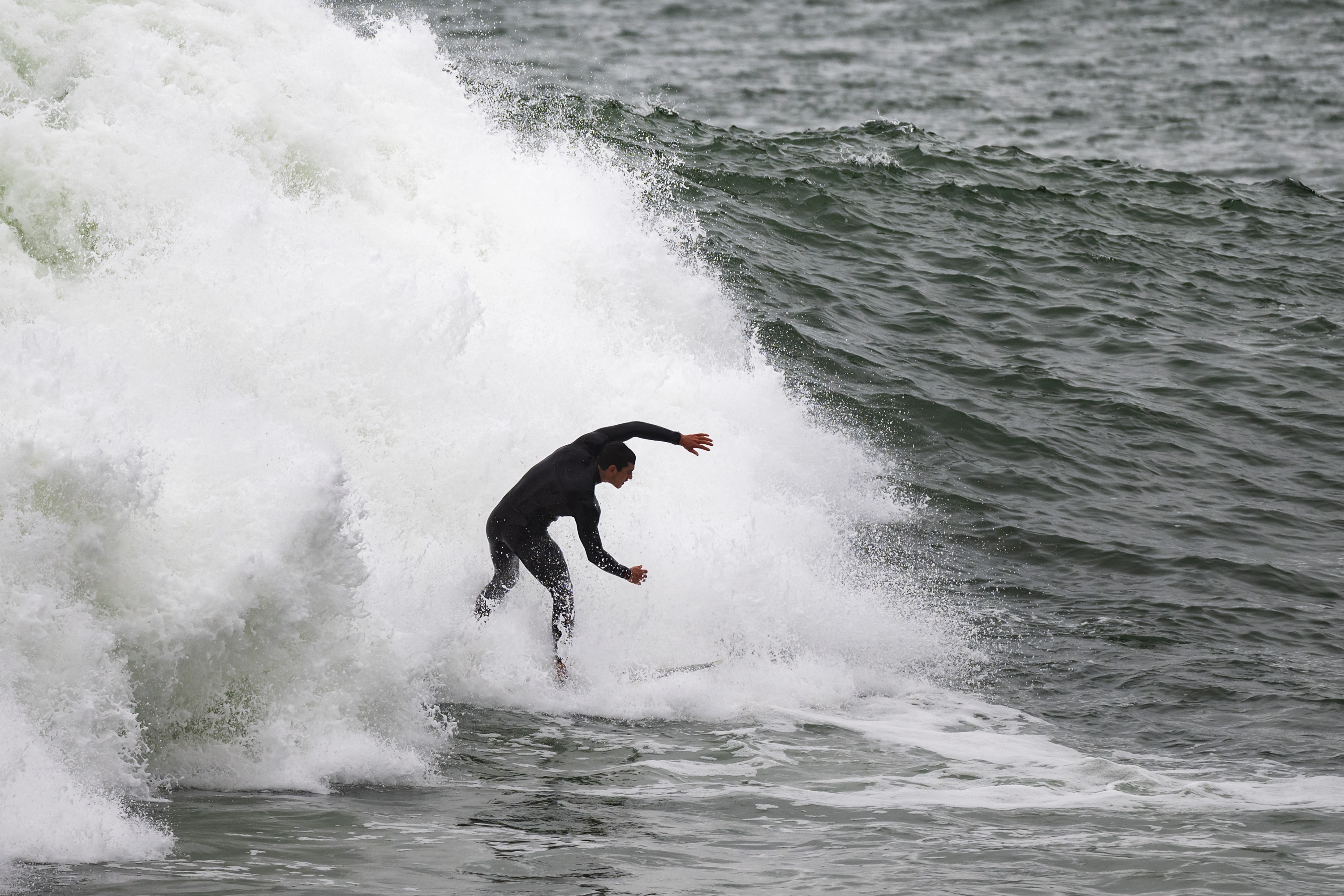 A surfer in a black wetsuit rides a powerful, frothy wave in the ocean, with water splashing dramatically around them.