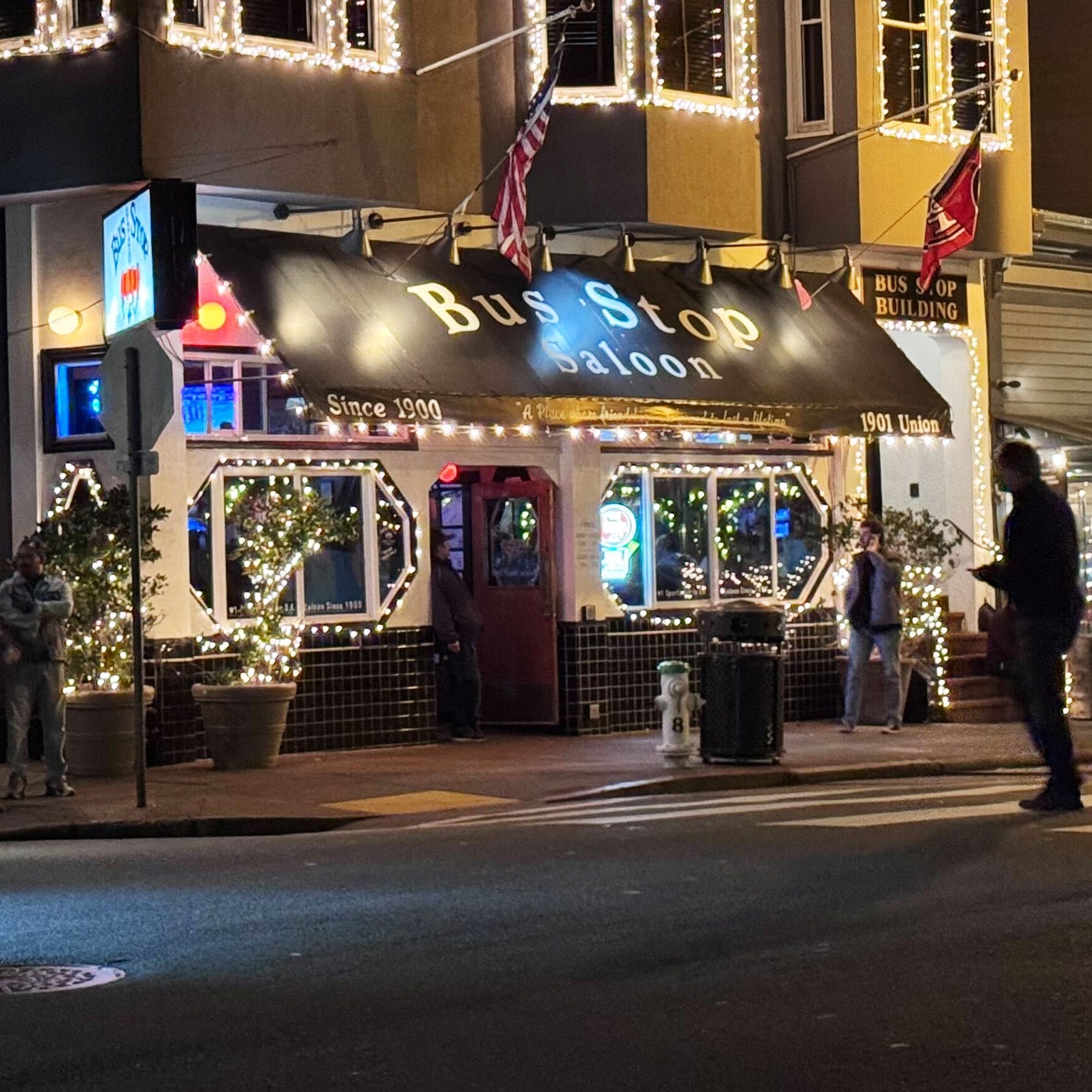 The image shows a warmly lit saloon named "Bus Stop" decorated with string lights. People are walking by on a crosswalk in front of the building at night.