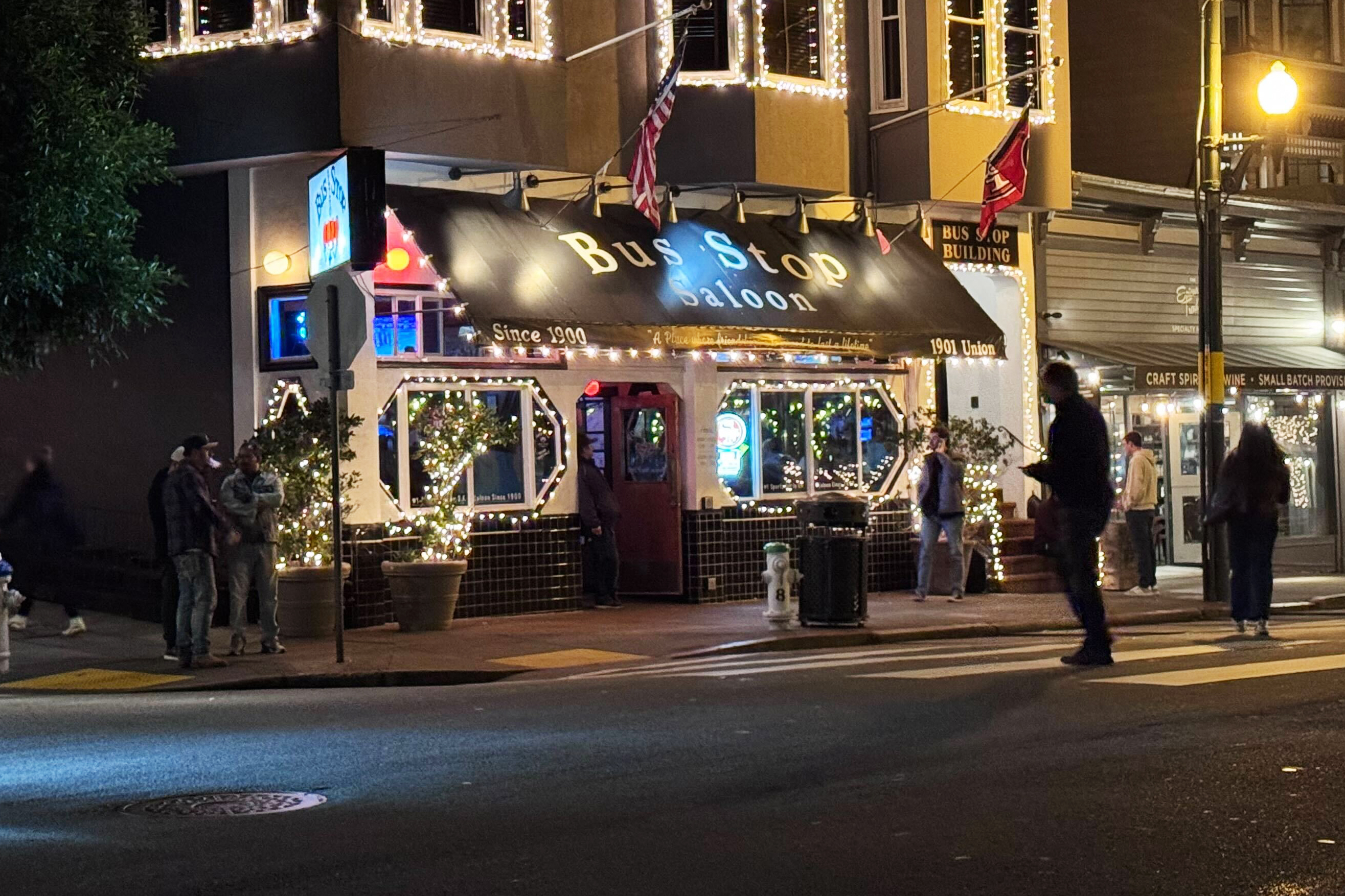 The image shows a warmly lit saloon named "Bus Stop" decorated with string lights. People are walking by on a crosswalk in front of the building at night.