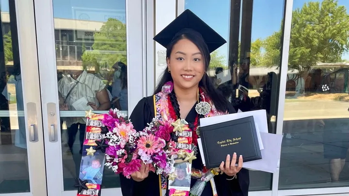 A graduate in cap and gown holds a diploma and a bouquet with colorful ribbons. She stands outside a building with glass doors.