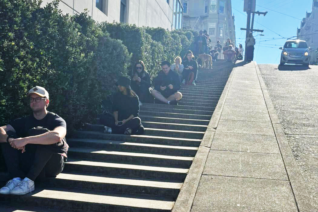 People are sitting on steep outdoor steps in a line, with greenery on one side. The scene is sunny, and a car is visible at the top of the hill.