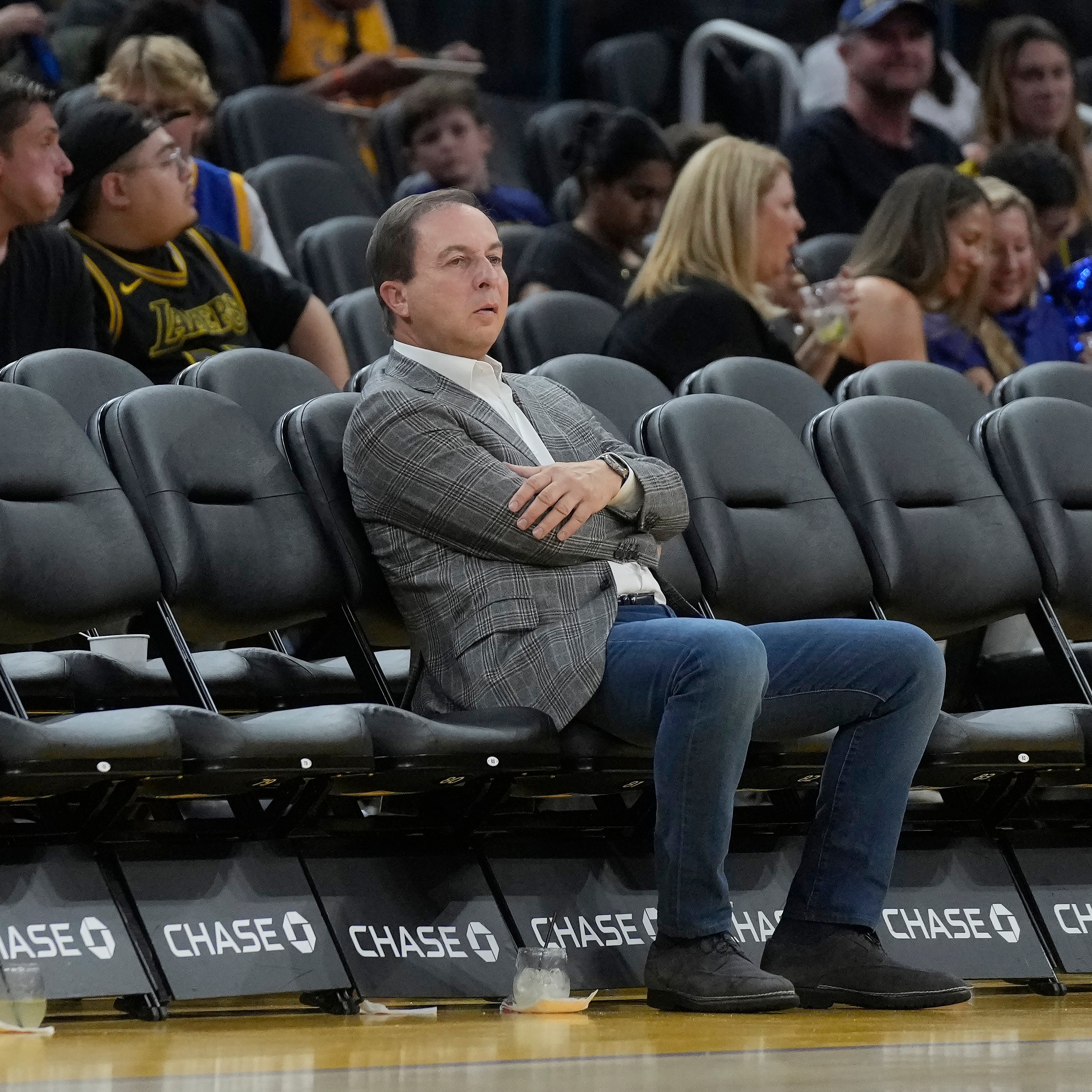 A man in a gray suit and crossed arms sits pensively on an empty row of black seats at a sports event, with a lively crowd behind him.