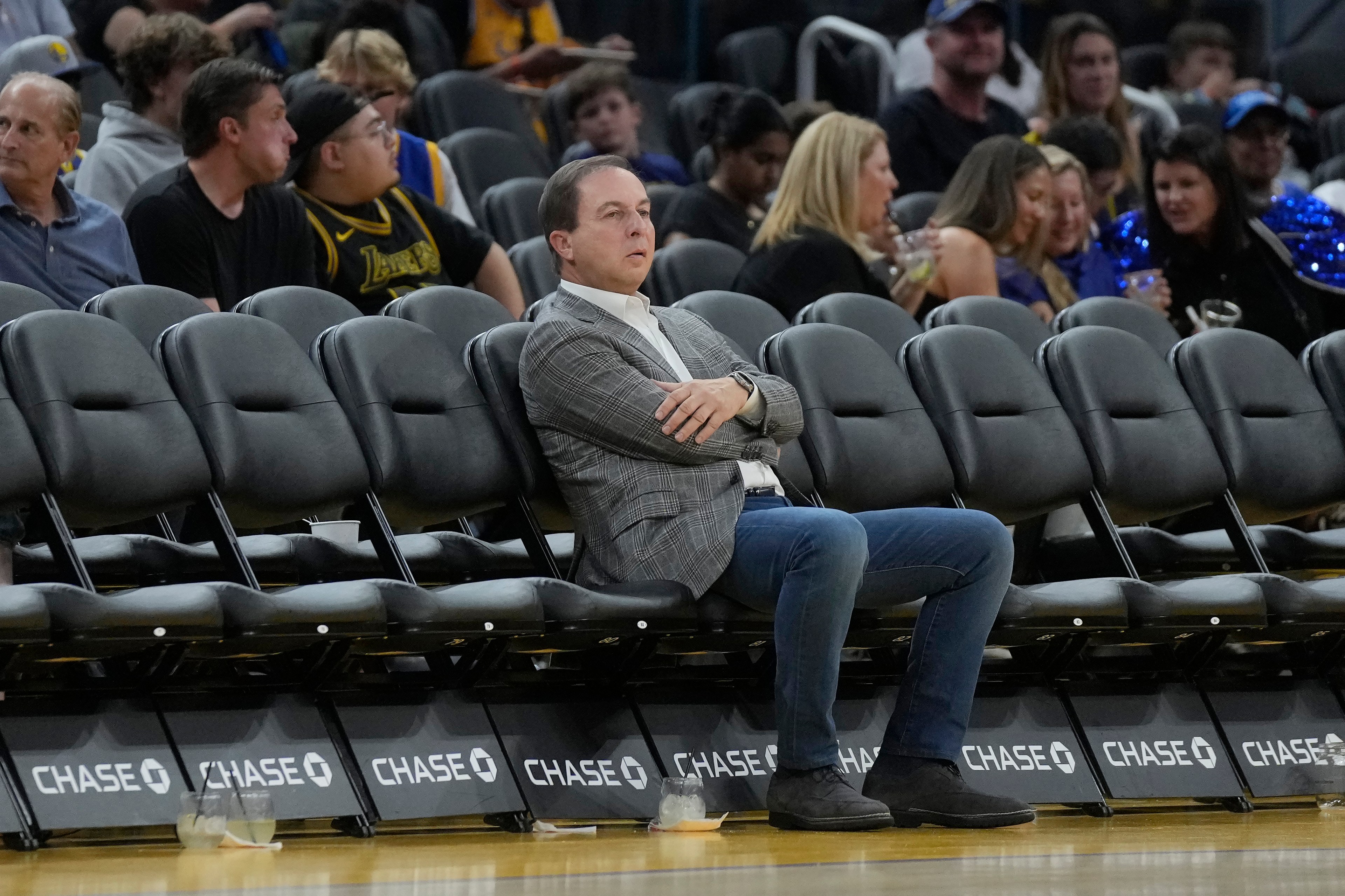 A man in a gray suit and crossed arms sits pensively on an empty row of black seats at a sports event, with a lively crowd behind him.