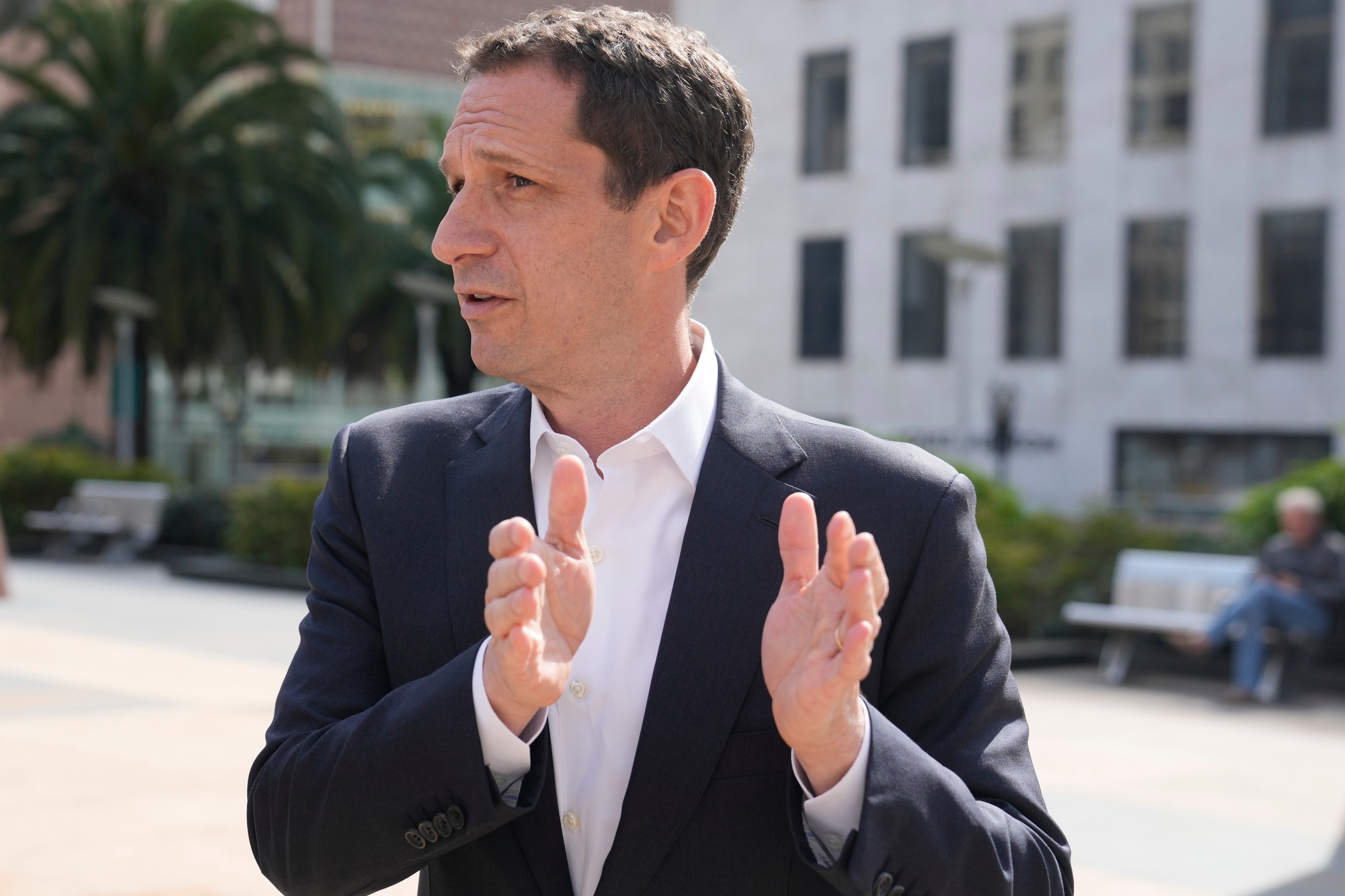 A person in a suit gestures outdoors in a city setting with palm trees and buildings in the background, conveying a sense of discussion or explanation.