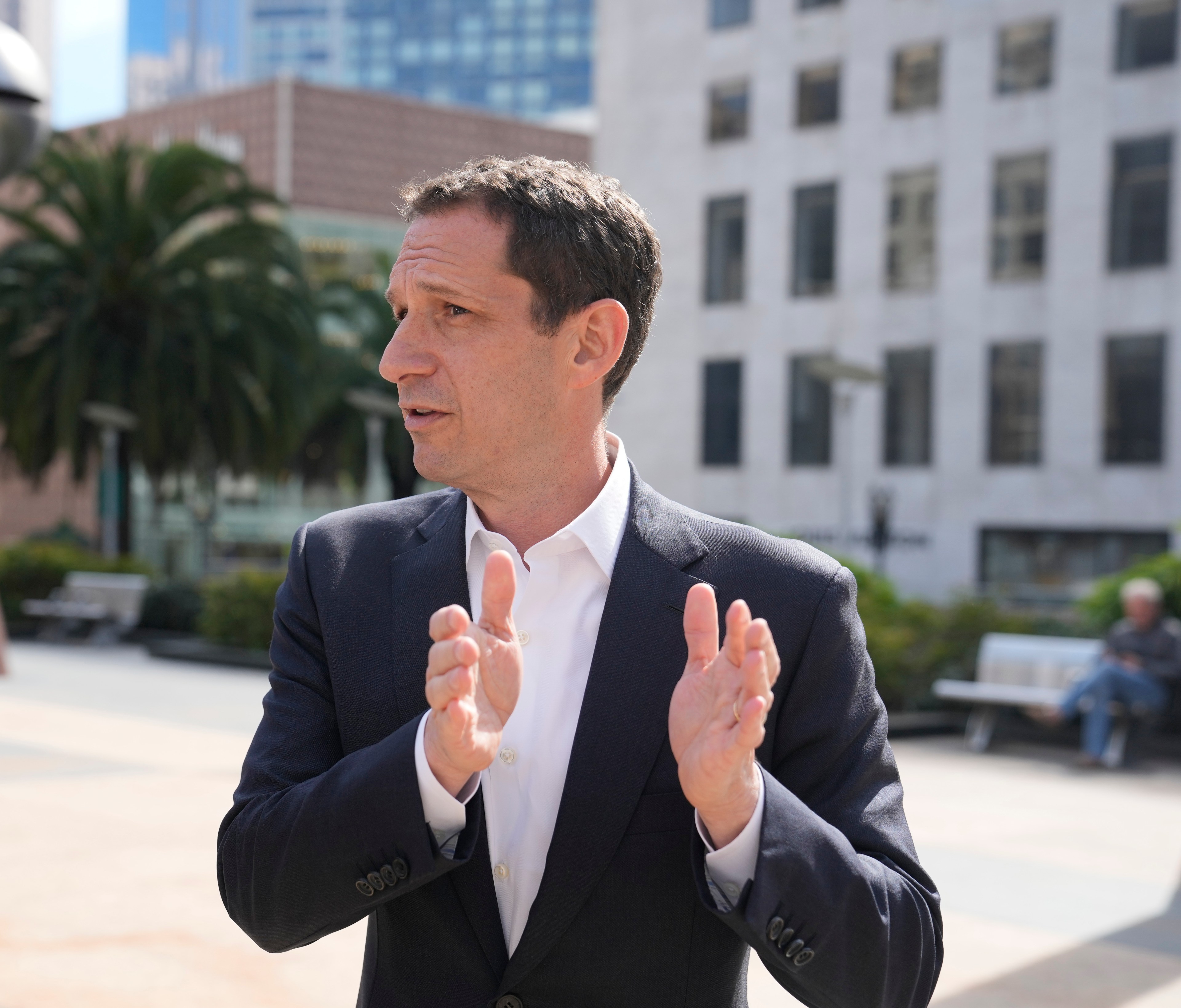 A person in a suit gestures outdoors in a city setting with palm trees and buildings in the background, conveying a sense of discussion or explanation.