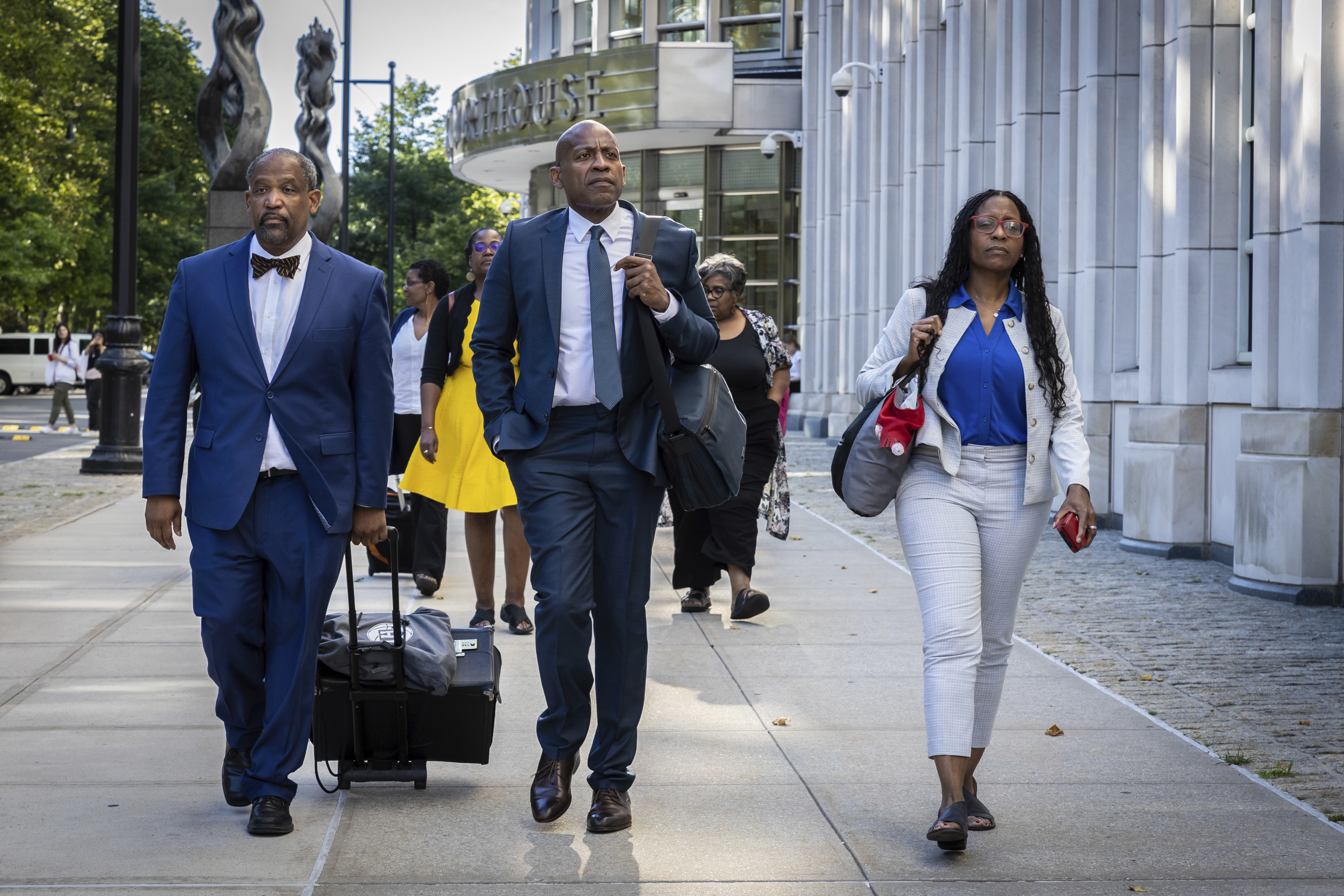 Three people are walking on a sidewalk; two men in suits and a woman in a white outfit. One man pulls luggage. A building and trees are in the background.