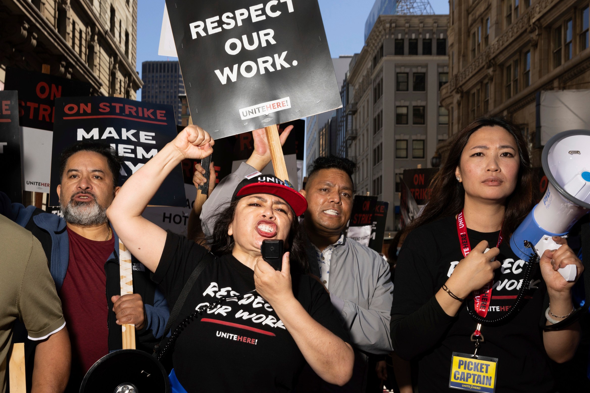 A group of people are passionately protesting in a city street, holding signs that read "RESPECT OUR WORK" and "ON STRIKE." One person speaks into a megaphone.