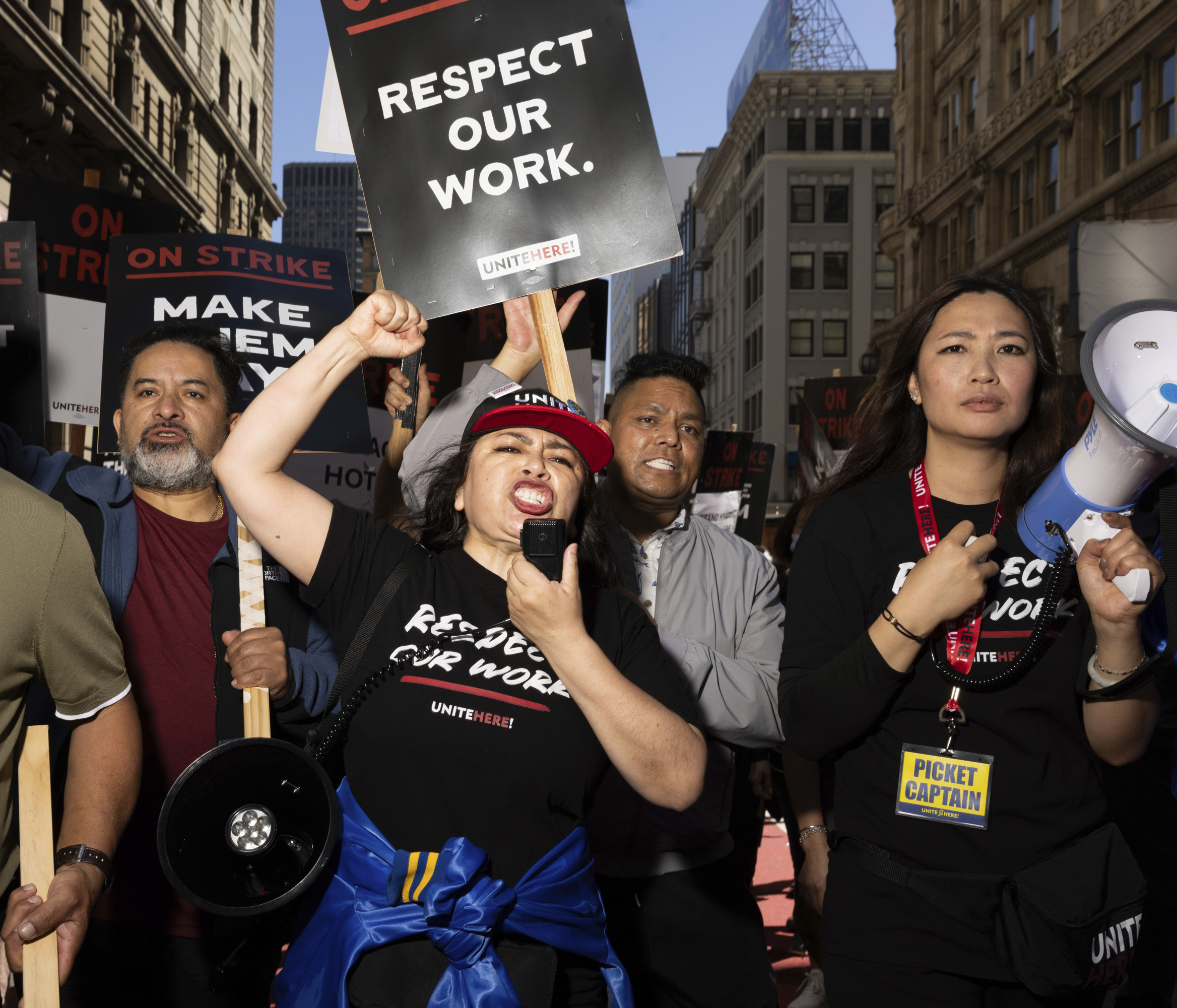 A group of people are passionately protesting in a city street, holding signs that read "RESPECT OUR WORK" and "ON STRIKE." One person speaks into a megaphone.