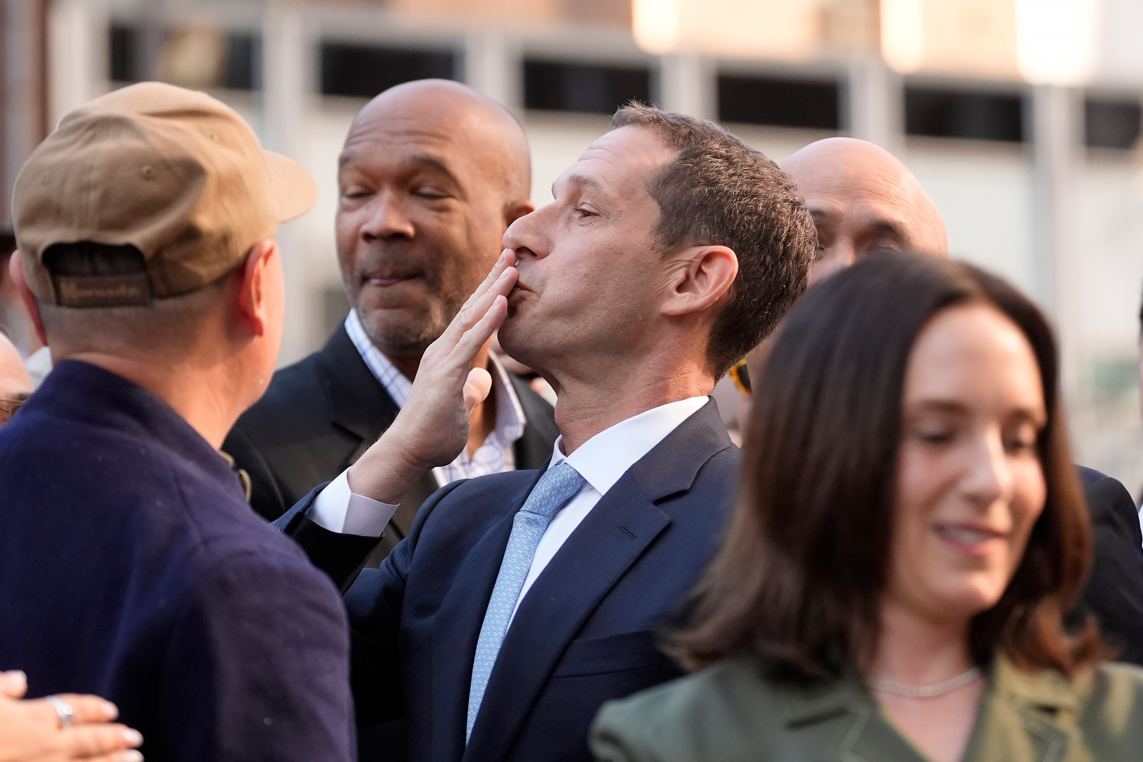 A man in a suit blows a kiss, surrounded by others in a crowded outdoor setting. He faces a person in a hat, with several people partially visible around them.