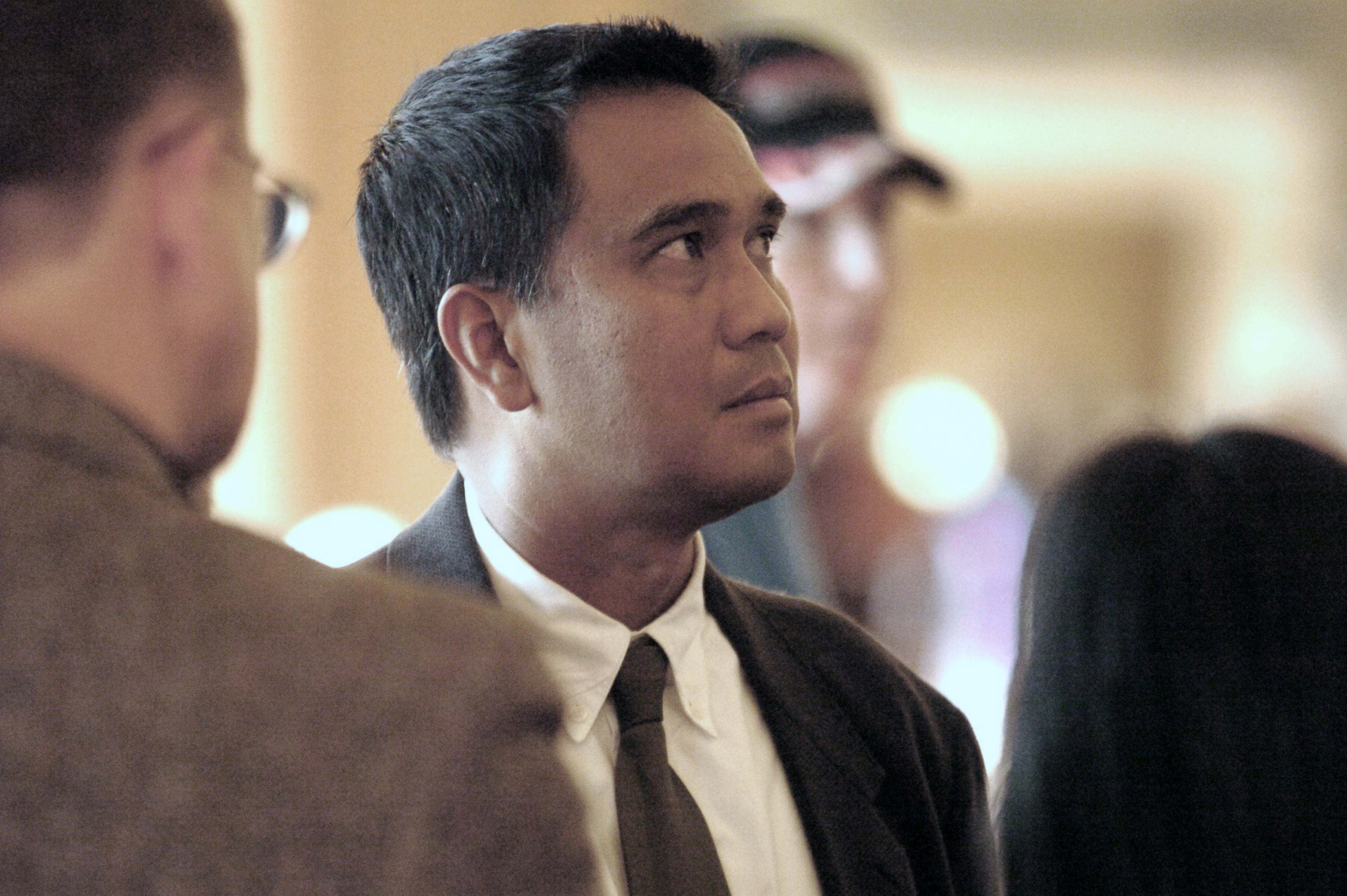 A man in formal attire with a focused expression looks upwards in a softly lit room. The blurred background suggests other people nearby.