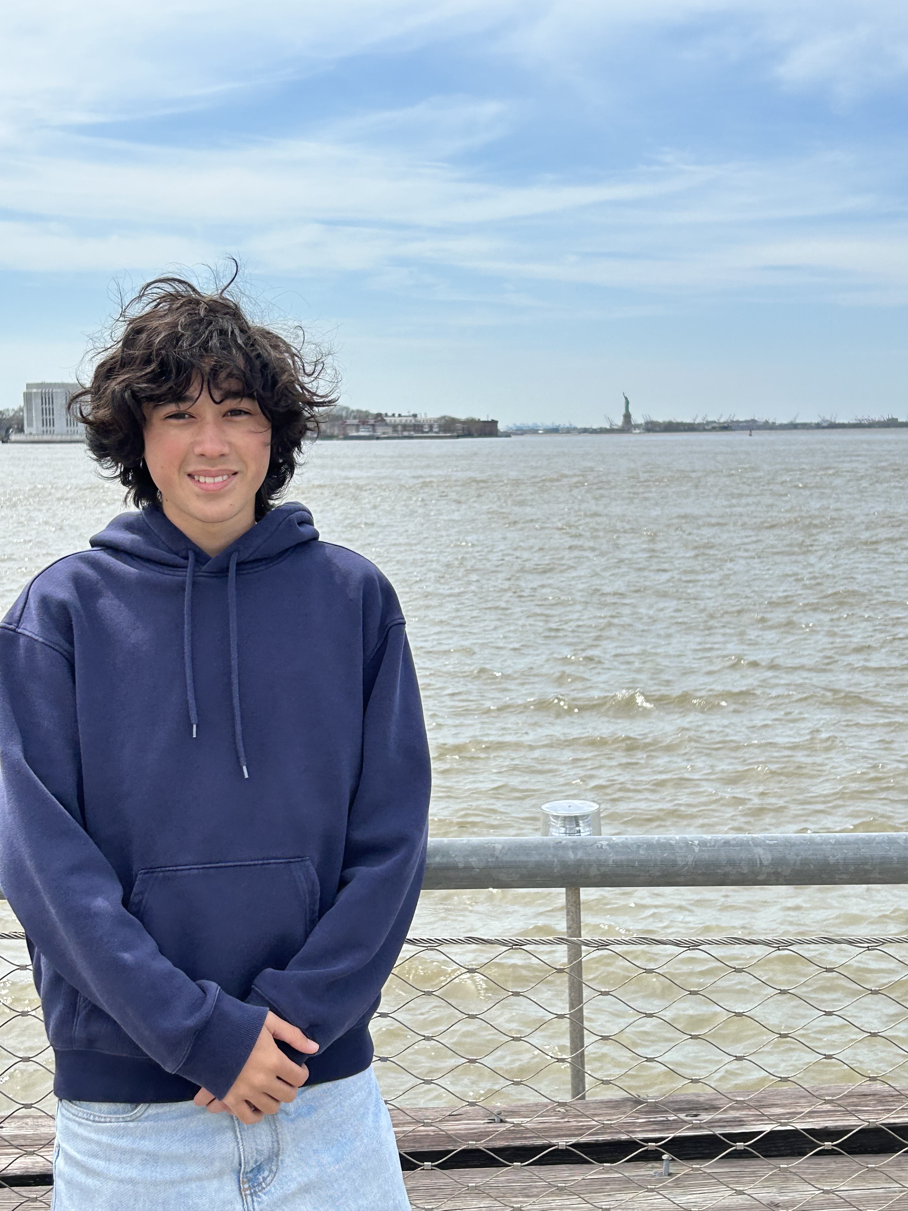 A person with curly hair in a navy hoodie stands by a railing near a body of water. The distant skyline includes the Statue of Liberty under a blue sky.