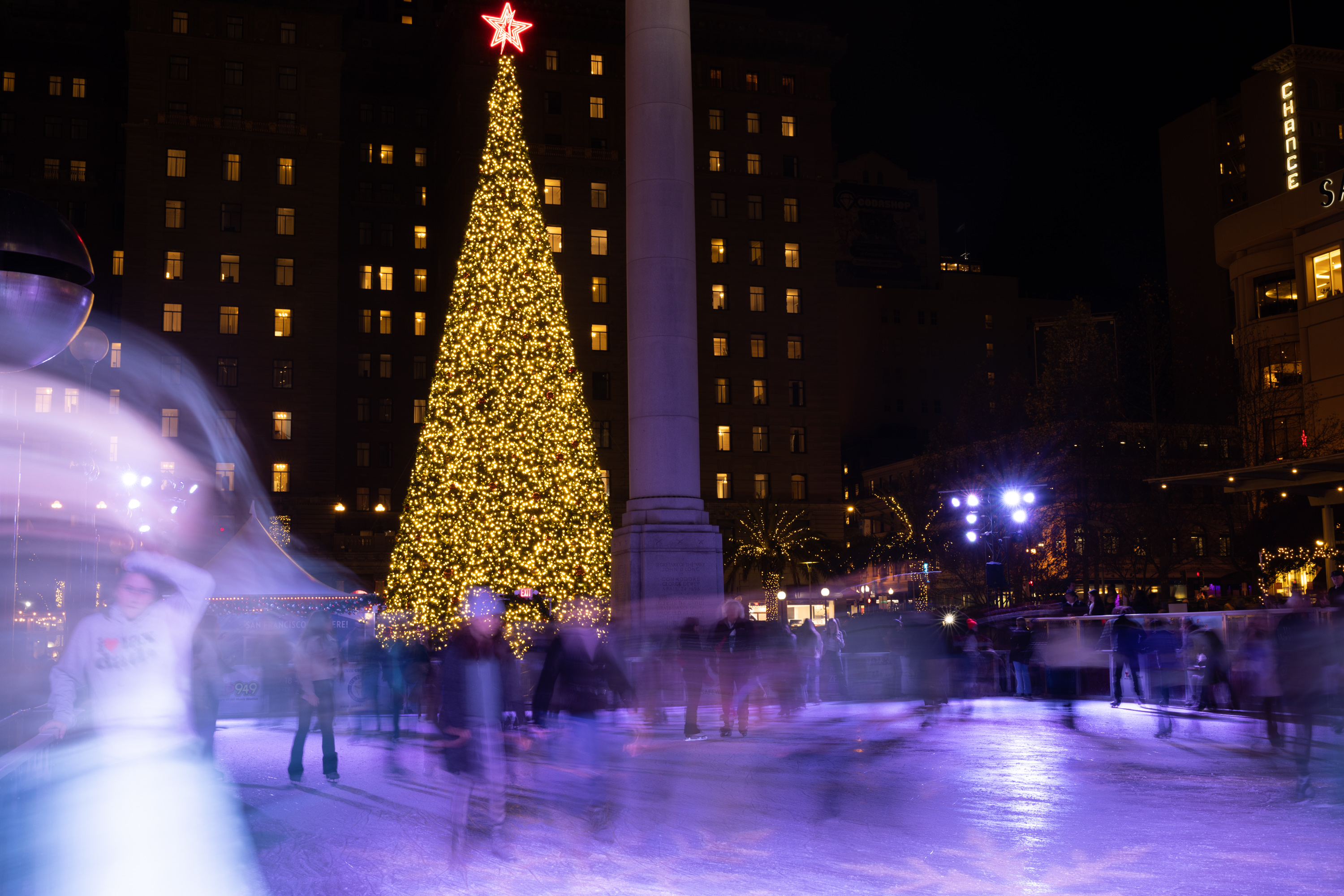 A large Christmas tree adorned with glowing lights stands prominently in a city square, surrounded by people ice skating under the night sky.