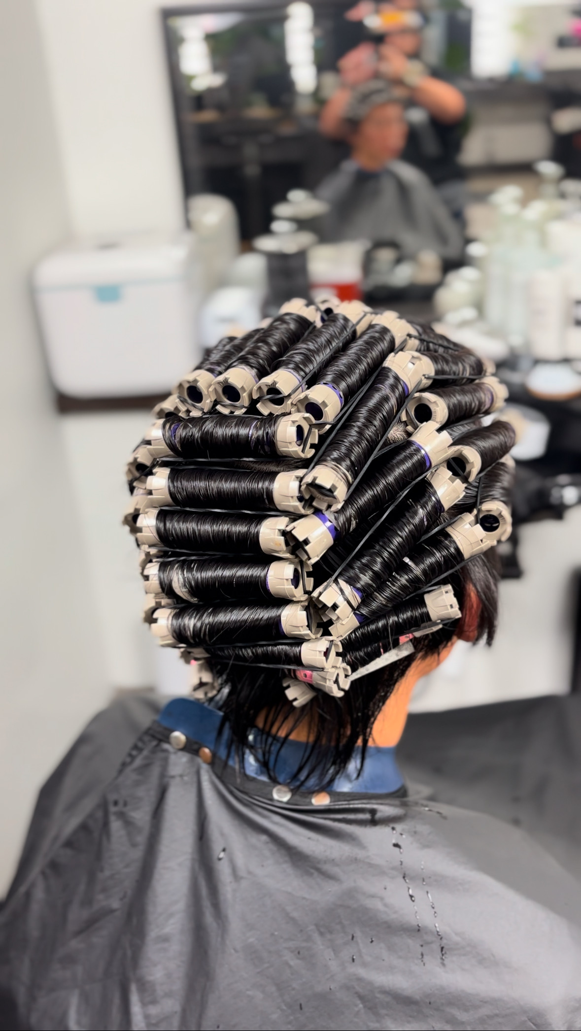 A person is having their hair set with numerous curlers, tightly wound for a perm, while sitting in a salon chair covered by a black cape.