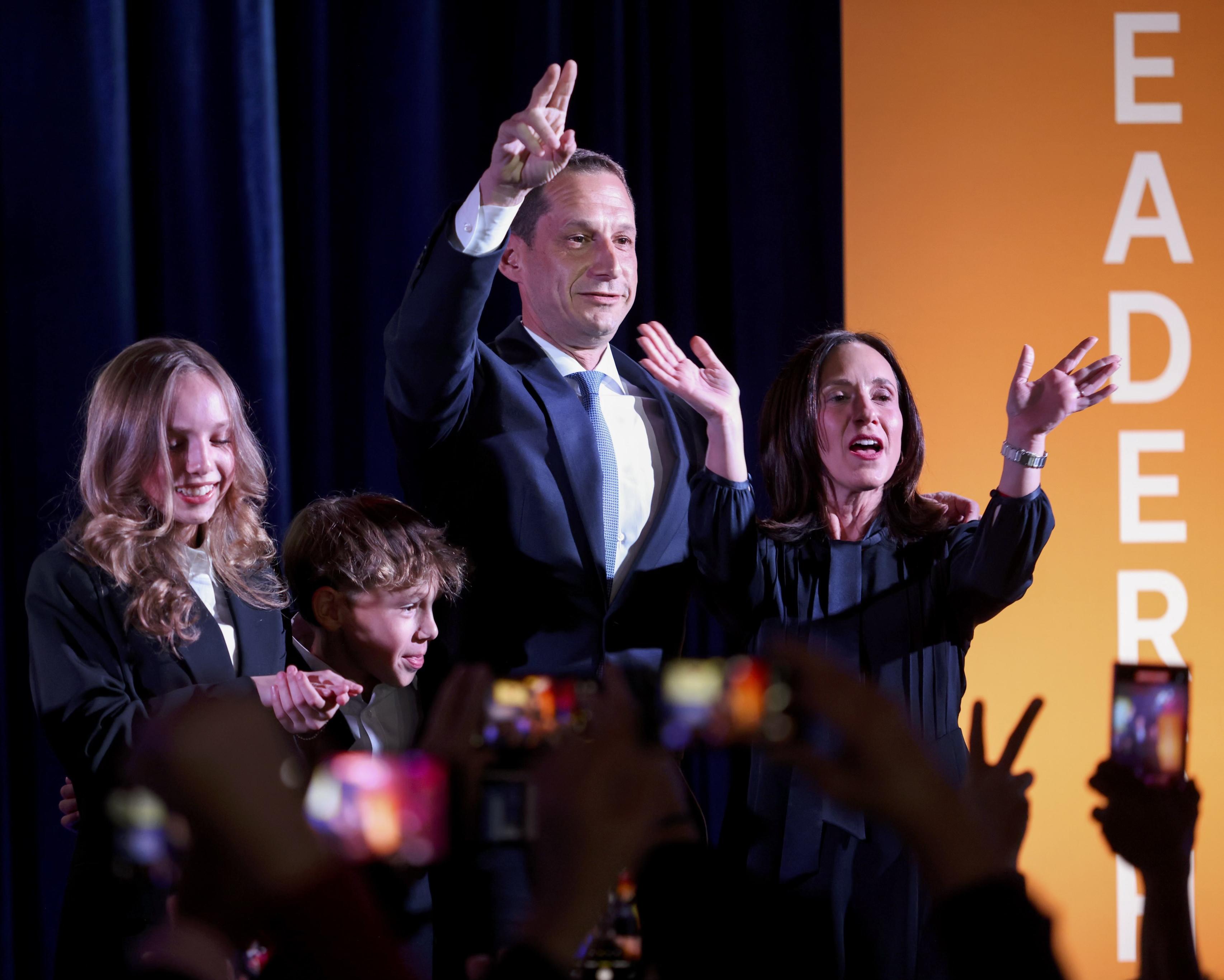 A man in a suit raises two fingers while standing with a woman and two children on stage, surrounded by a cheering crowd against a dark background with orange text.