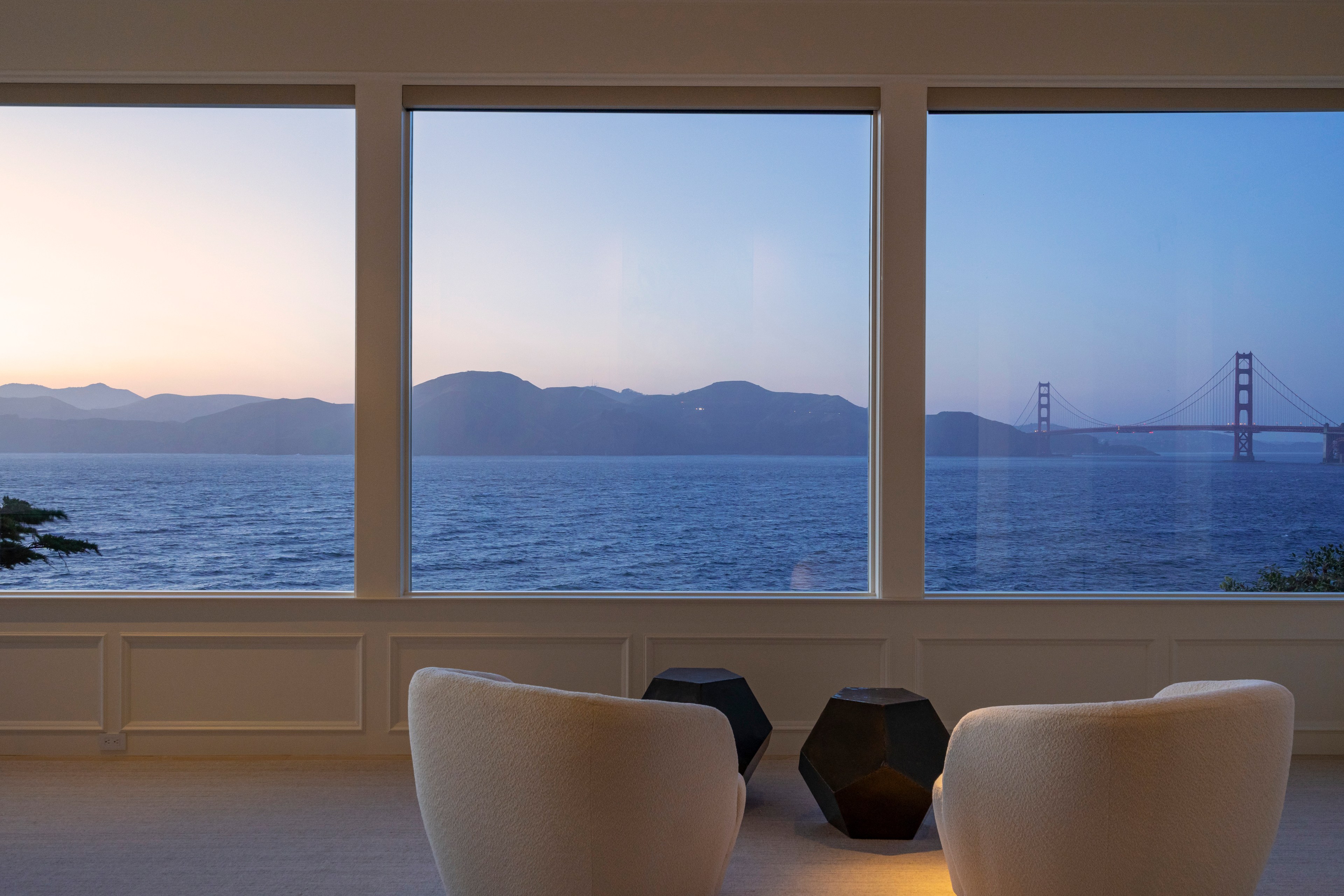 Two white chairs face large windows with a view of a calm sea and distant mountains. A bridge is visible to the right against a clear evening sky.