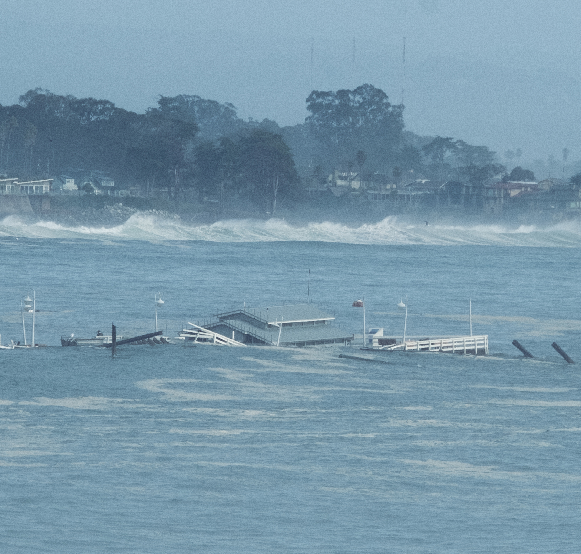 A partially submerged pier with scattered debris is visible in rough ocean waters, with waves crashing closer to a shoreline lined with trees and buildings.