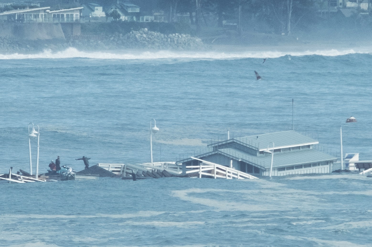 A large structure, partially submerged, floats amid turbulent water. Nearby, people stand on a dock, with waves crashing in the background, under a cloudy sky.