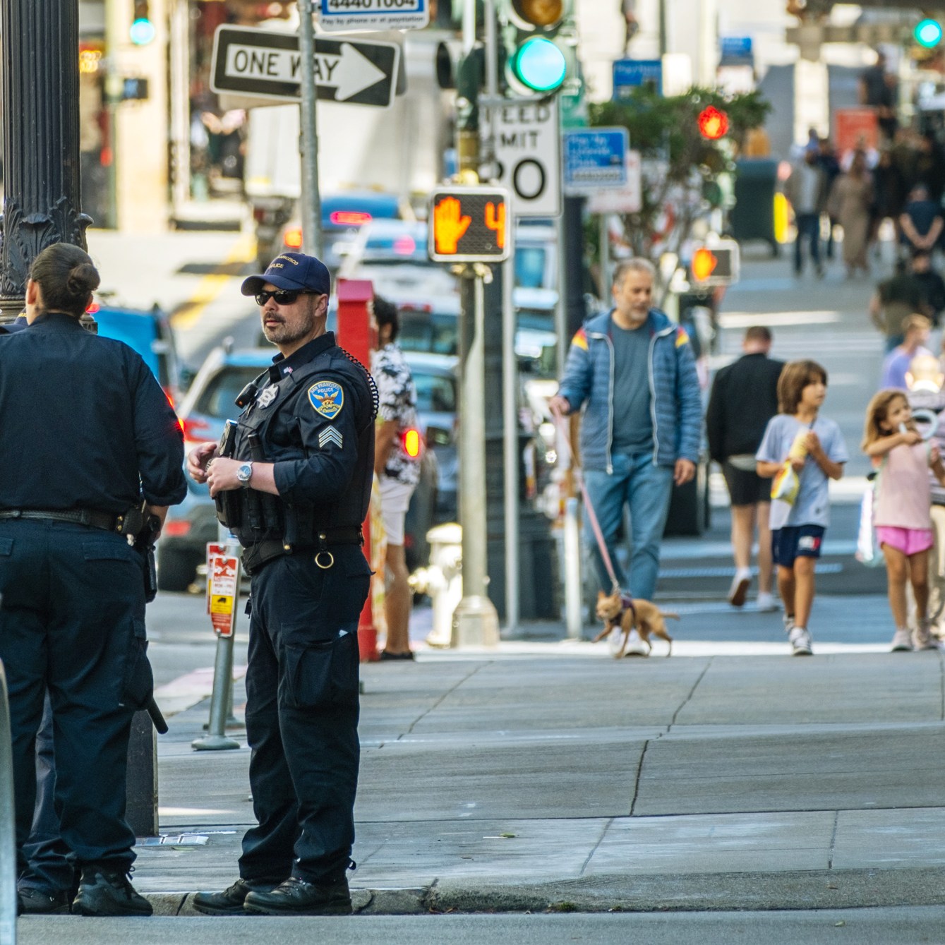 Police officers stand by a vehicle on a city street, while pedestrians, including children and a person walking a dog, pass by on the sidewalk.