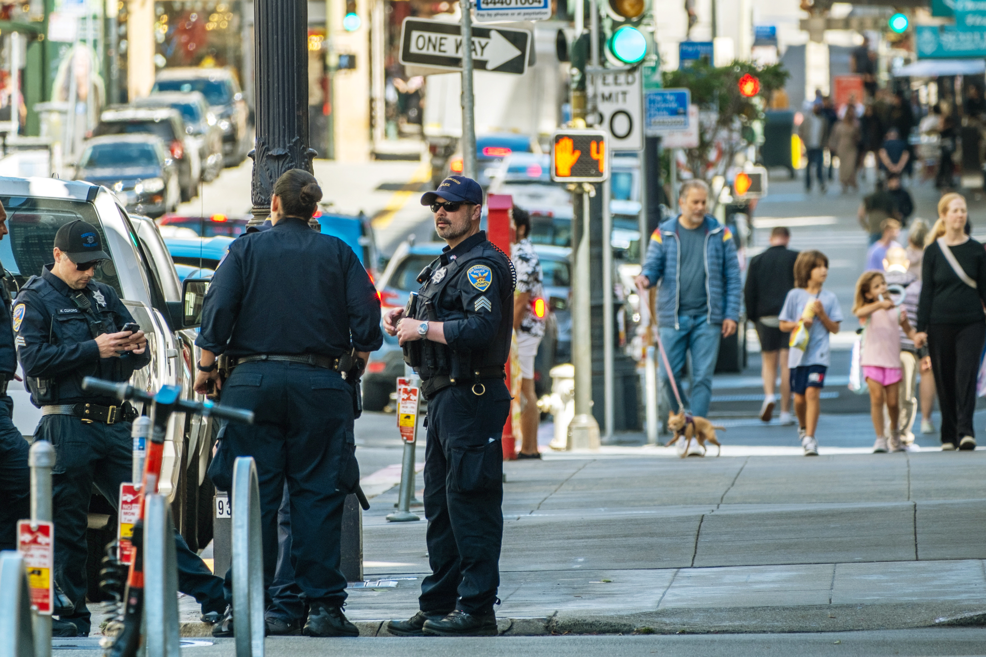 Police officers stand by a vehicle on a city street, while pedestrians, including children and a person walking a dog, pass by on the sidewalk.