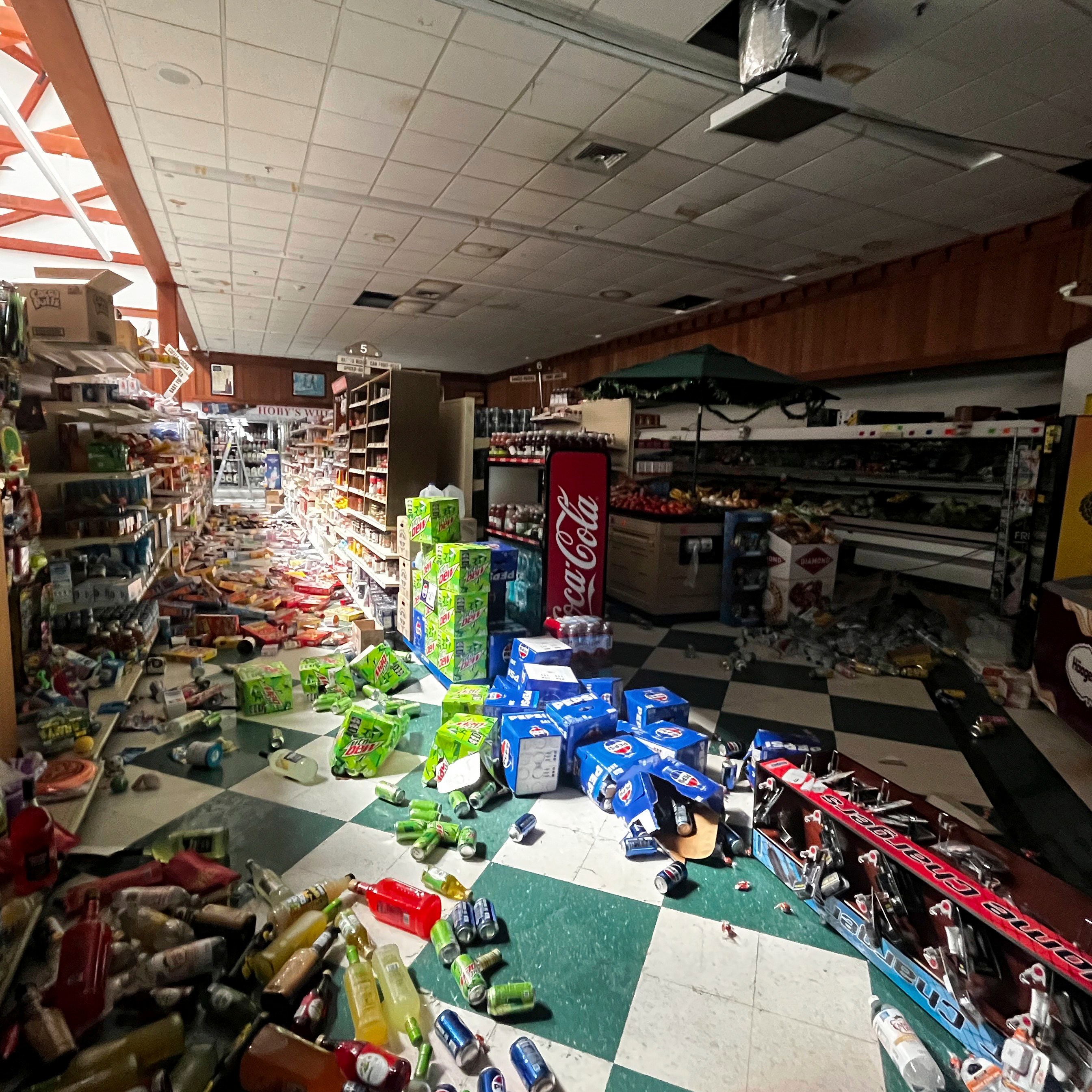 The image shows a store interior with products scattered across the floor, including knocked-over bottles and boxes. The ceiling appears damaged, and the scene is chaotic.