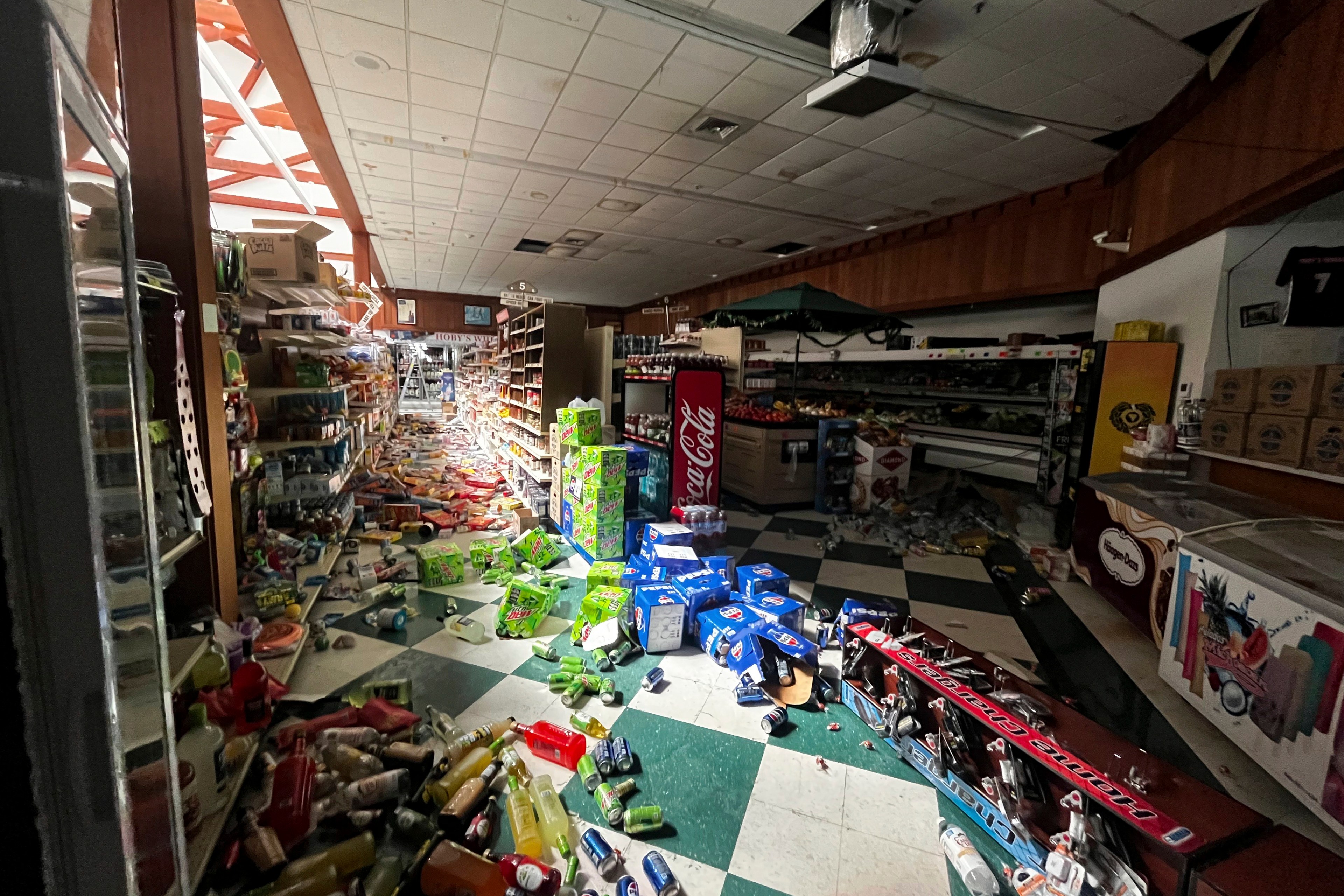 The image shows a store interior with products scattered across the floor, including knocked-over bottles and boxes. The ceiling appears damaged, and the scene is chaotic.