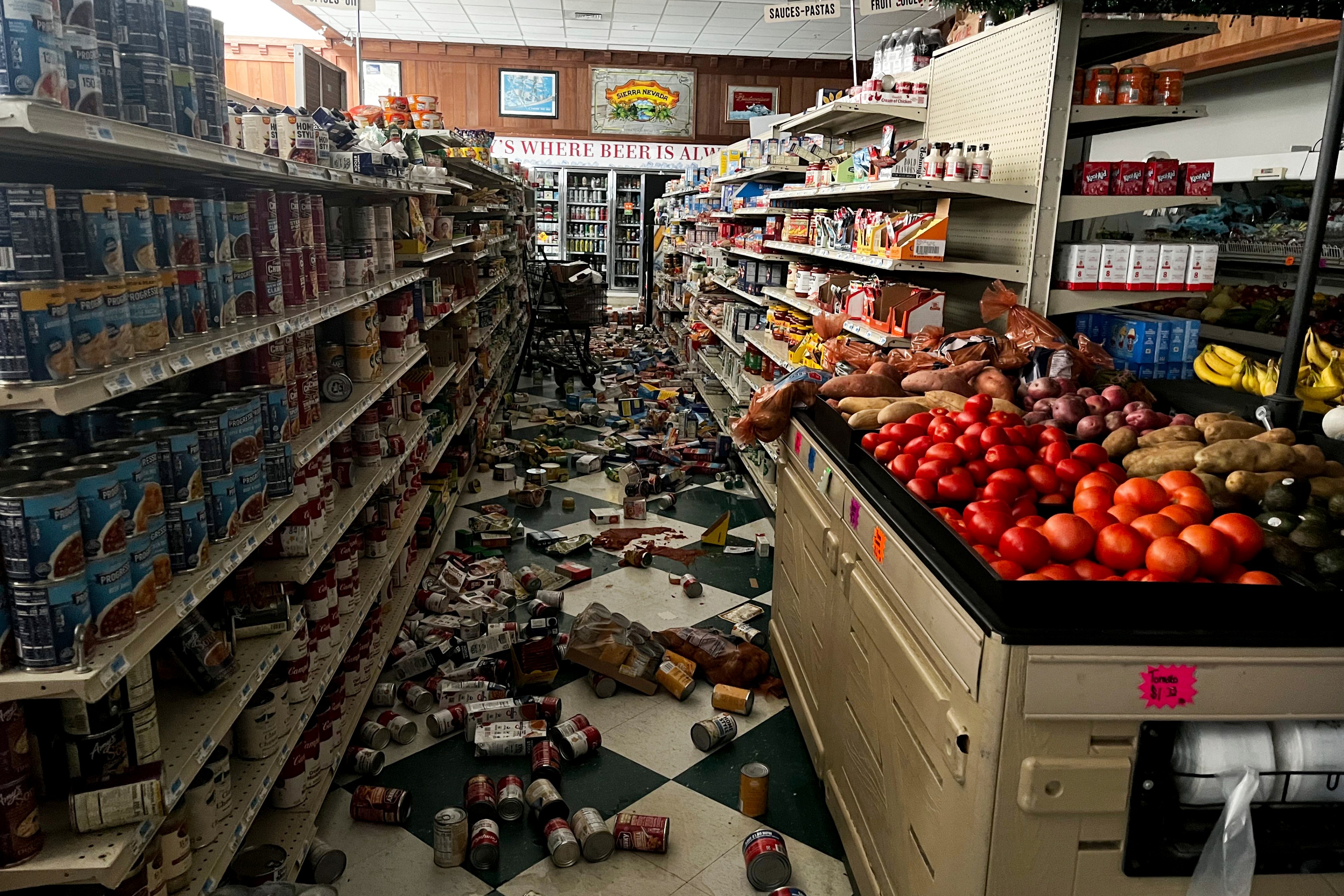 A disordered grocery aisle with cans and boxes scattered on the floor, surrounded by stocked shelves and a produce section with tomatoes and potatoes.