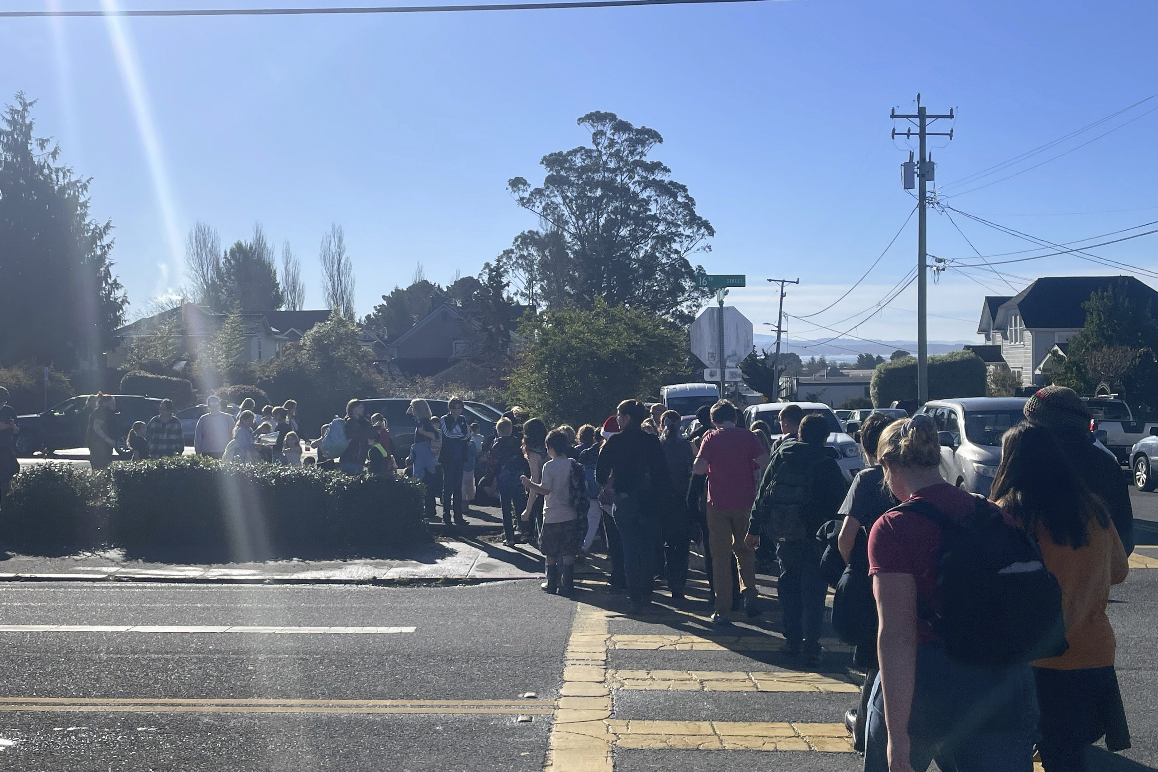 A group of people is gathered on a street corner near parked cars, under a clear blue sky. They appear to be waiting, with some facing away from the camera.