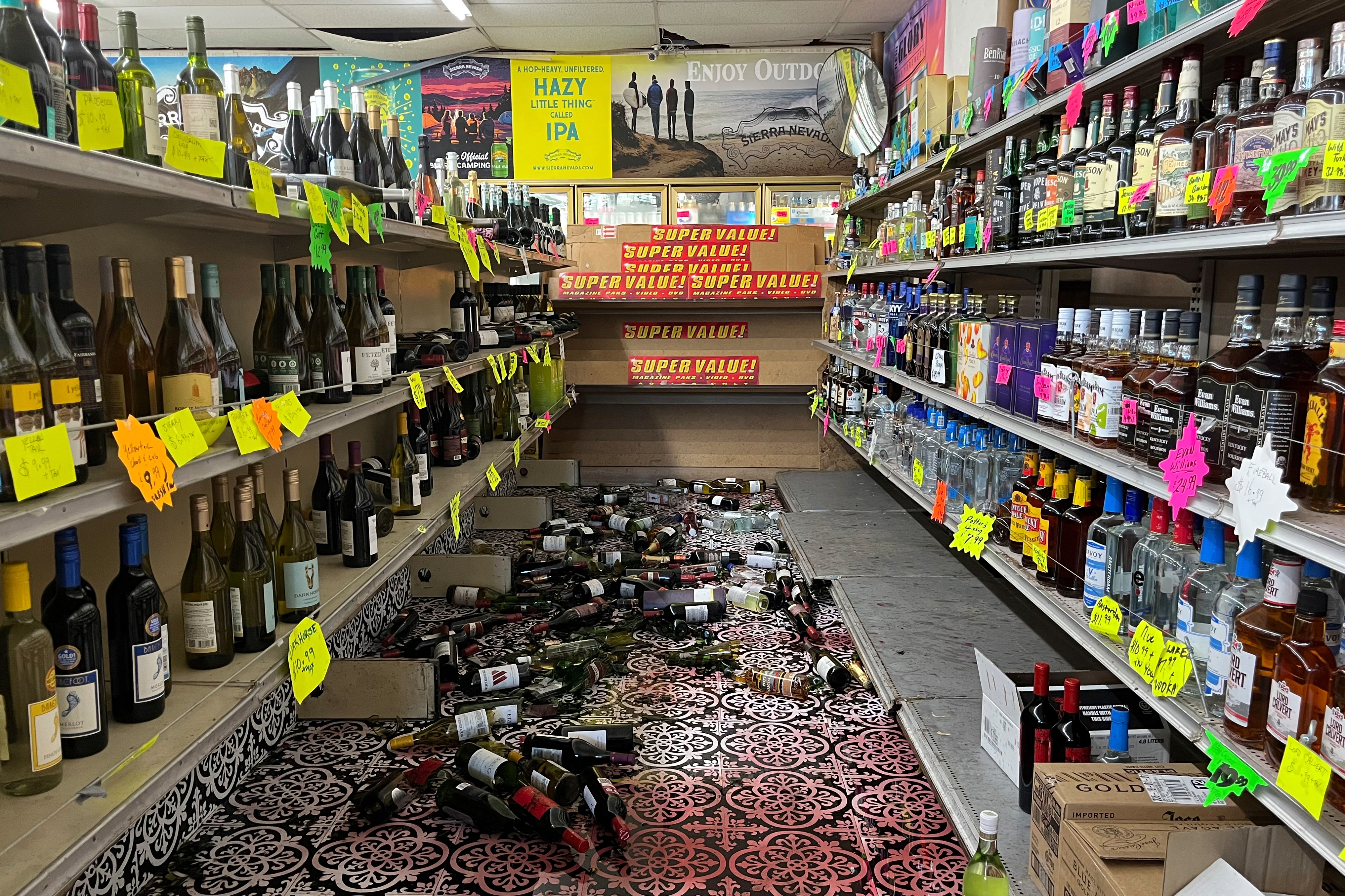 A liquor store aisle is shown with bottles toppled over and scattered on the floor, while shelves on both sides hold upright bottles with colorful price tags.