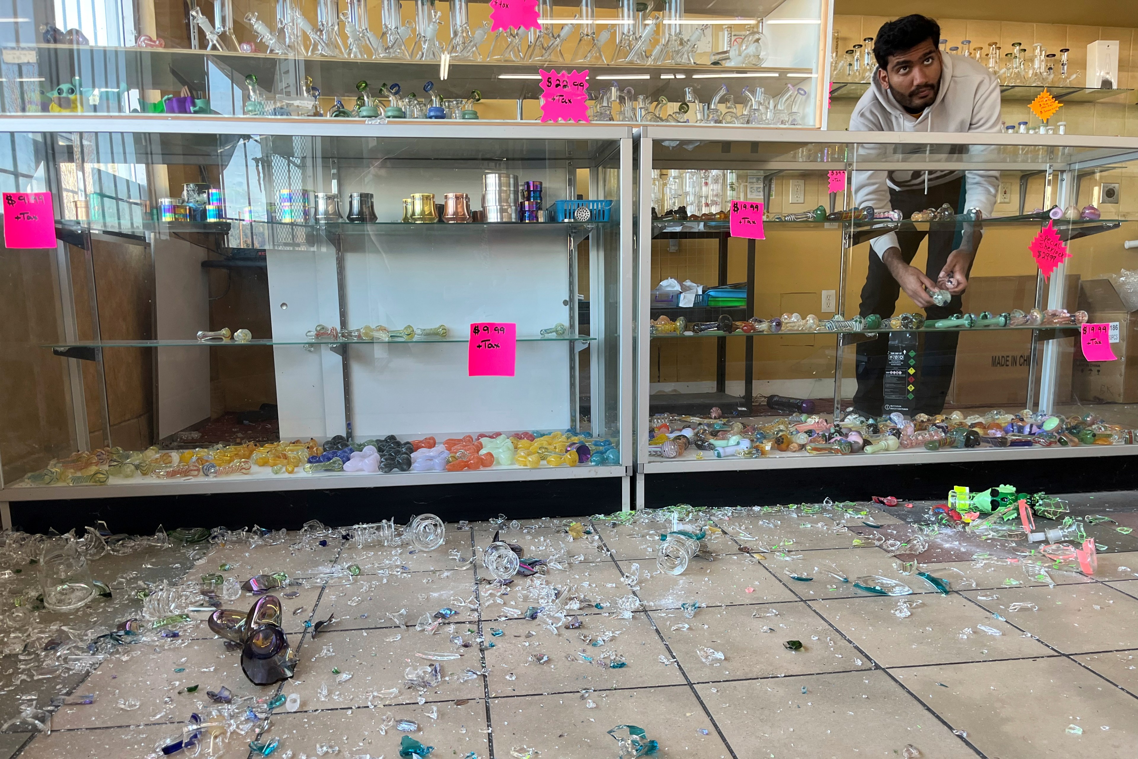 A man in a store gathers items from broken glass cases. Colorful glass pipes and pieces are scattered across the floor with bright signs on the cases.