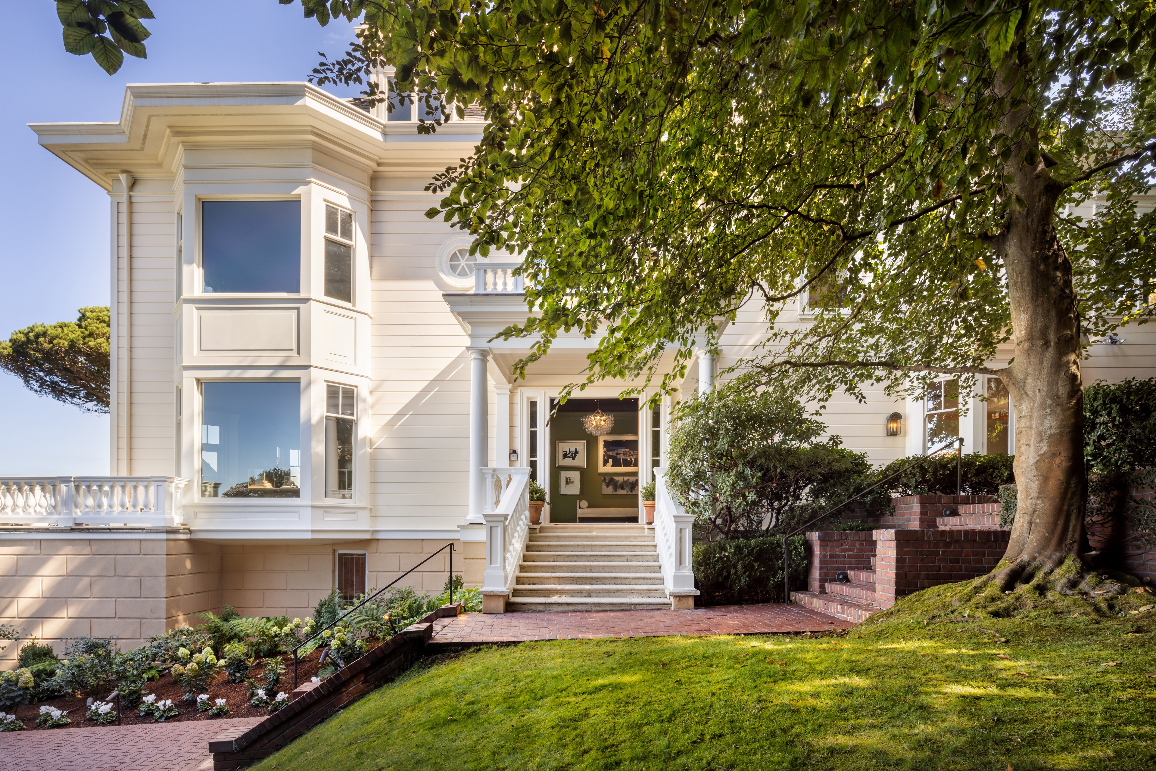 A white, two-story house with large windows and a porch is surrounded by greenery. Brick steps lead up to the entrance, framed by a tree and manicured lawn.