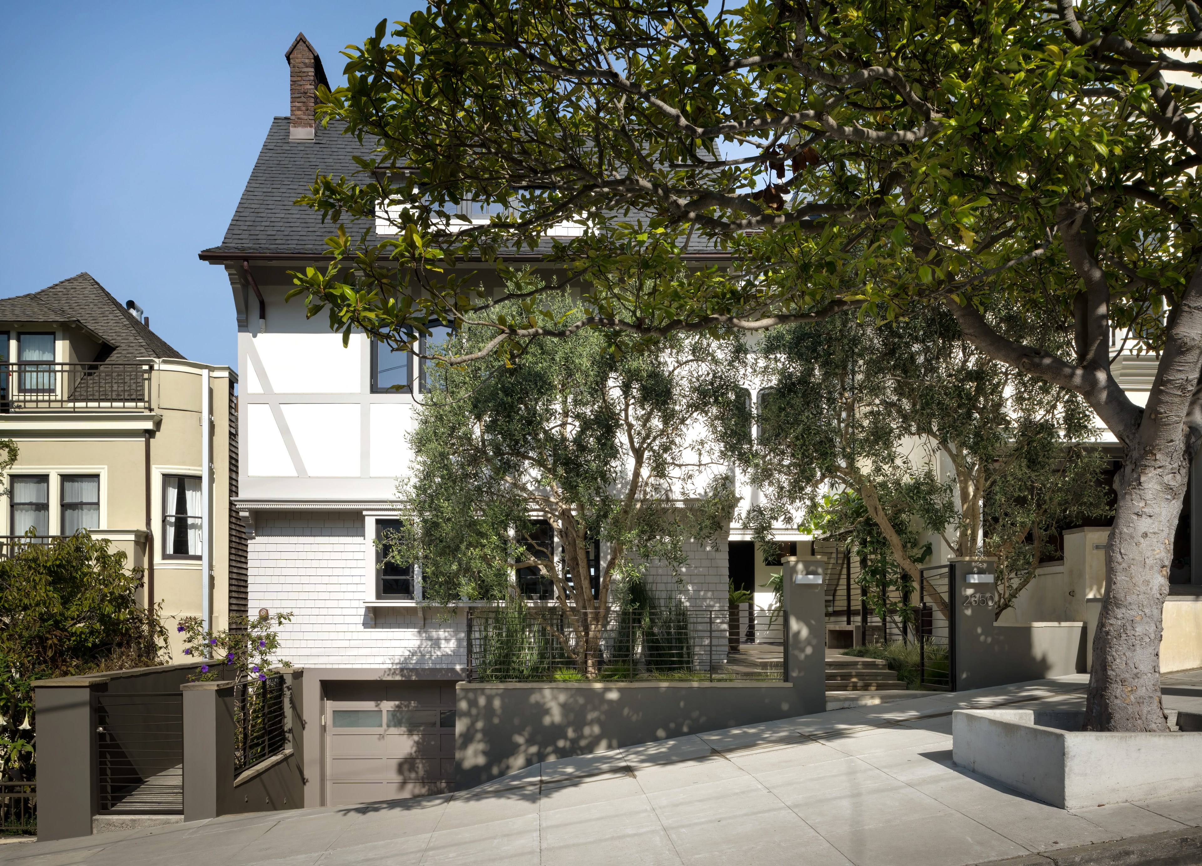 The image shows a modern two-story house with a steep roof and a white facade. Trees partially obscure the view, and it's situated on a sloped street.