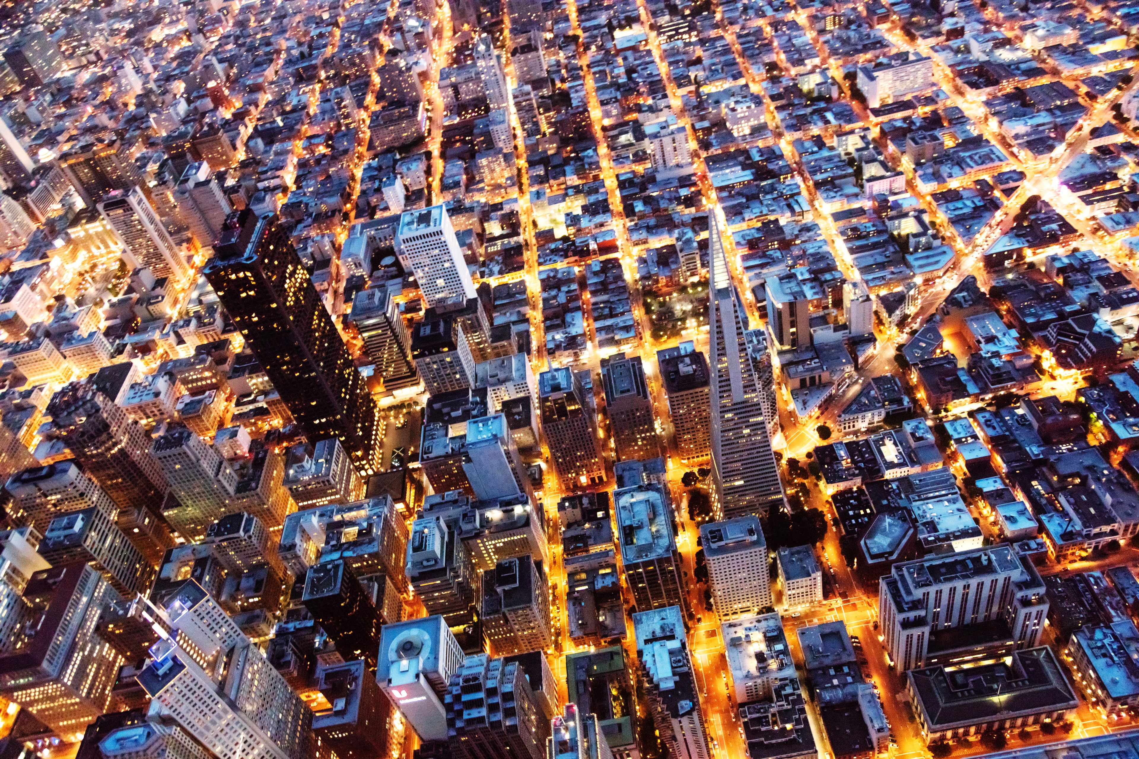 The image shows an aerial night view of a bustling city with a grid-like street layout. Tall buildings are illuminated, with glowing streets forming a vibrant pattern.