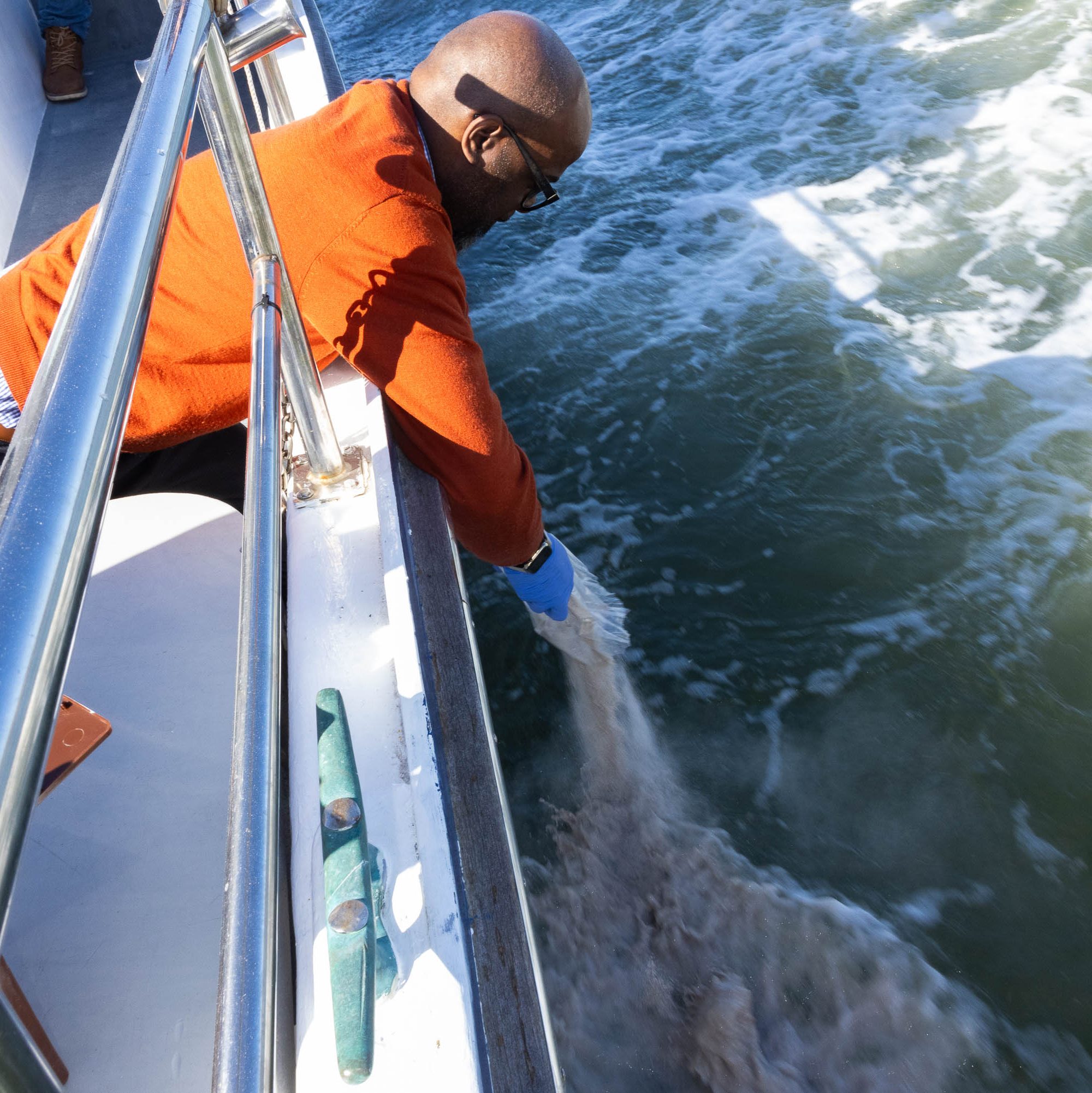 A man leans over the side of a boat, touching the water, with waves around. He drops ashes.