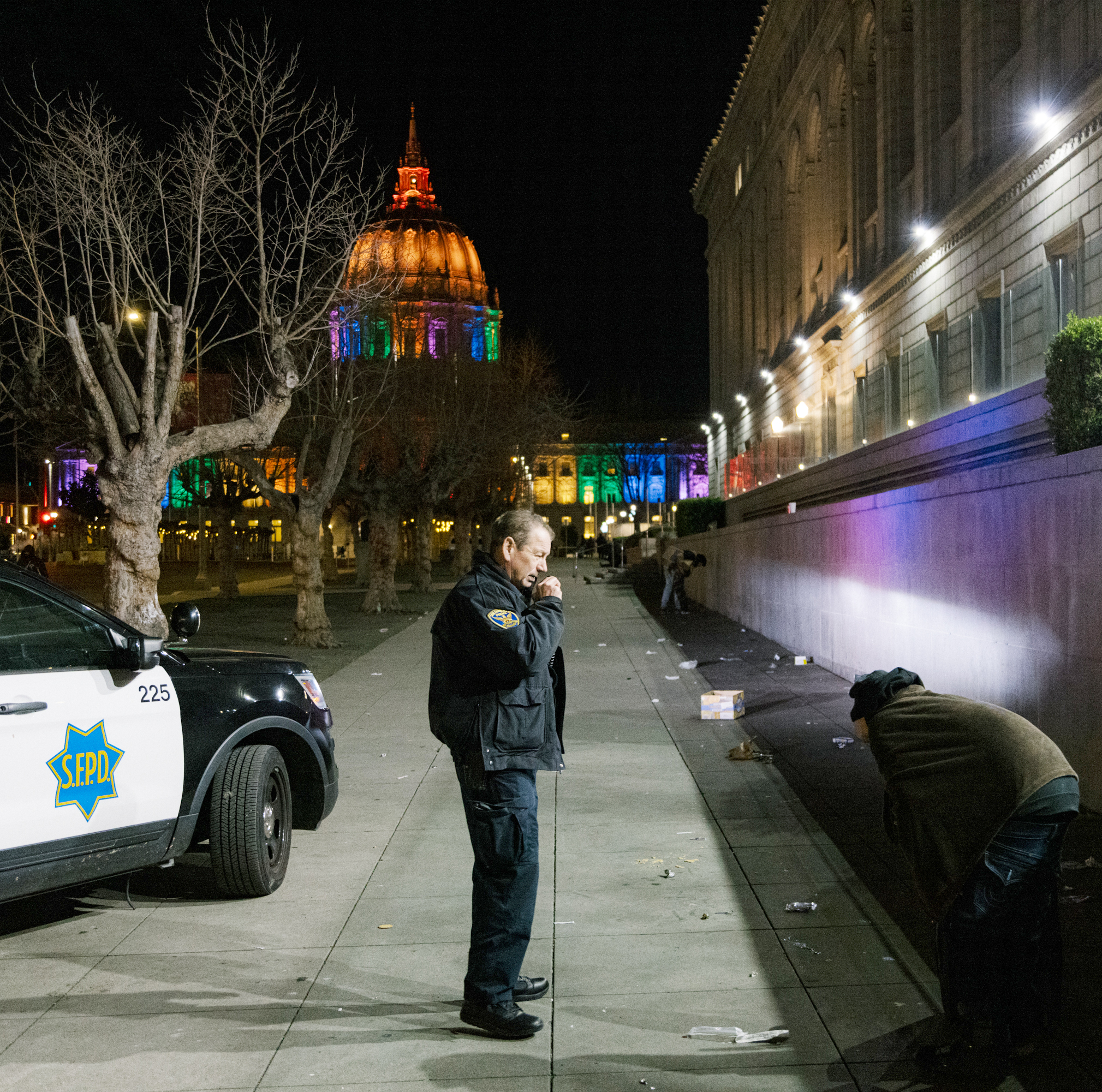 A police officer stands near a patrol car with &quot;SFPD&quot; on it, while a hooded person leans against a wall. In the background, a lit-up dome in rainbow colors is visible.