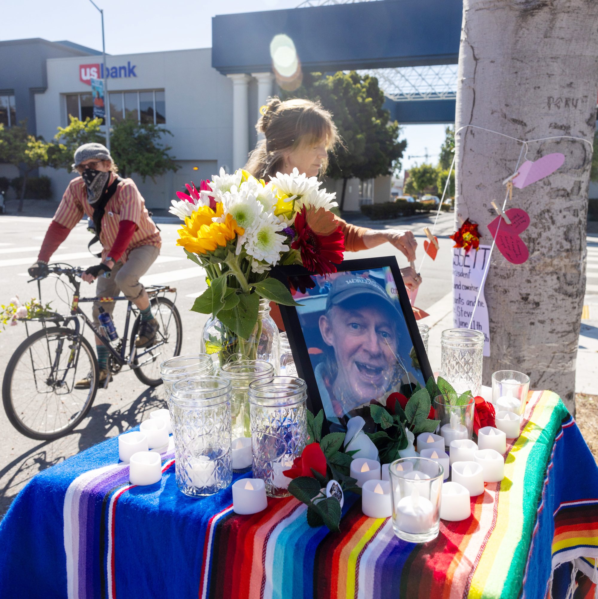 A memorial set on a city street with a photo, flowers, candles, and a colorful cloth atop a small table. A person bikes past.