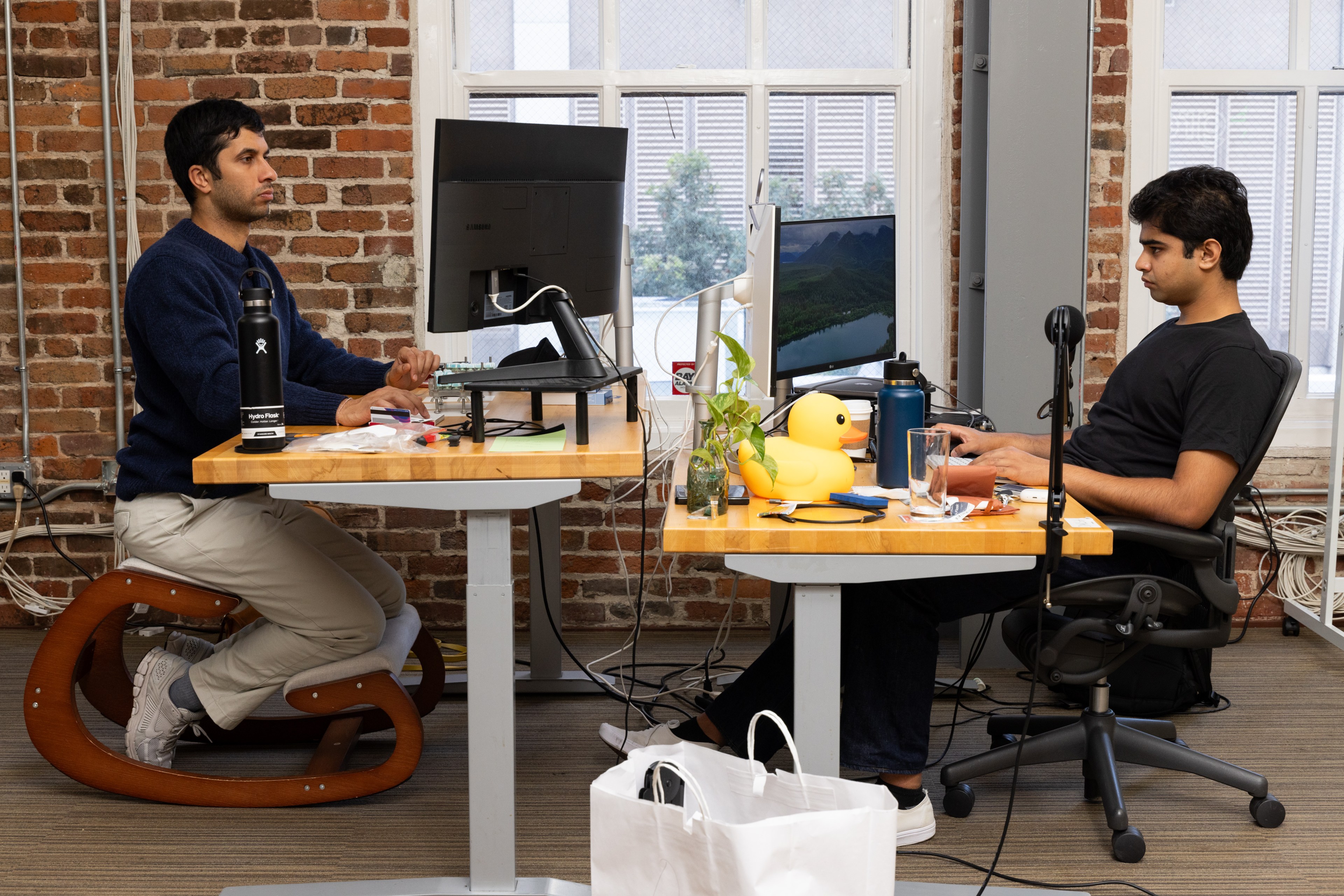 Two people are working at adjacent desks in an office. One sits on a kneeling chair; the other in a standard office chair. Monitors, mugs, and a rubber duck are on the desks.