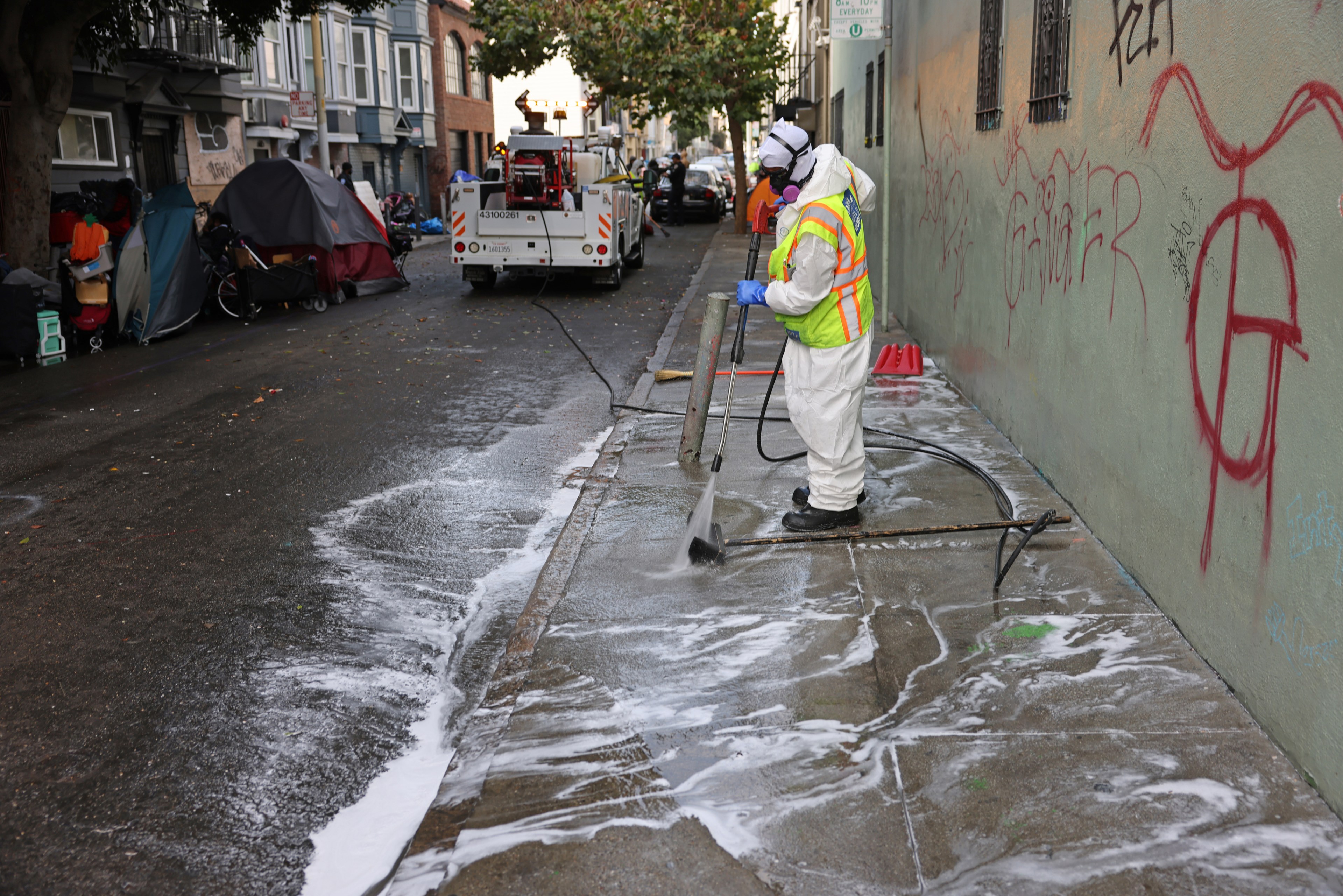 A worker in protective gear pressure washes a graffiti-covered sidewalk next to a row of tents, while a utility truck is parked nearby on the street.