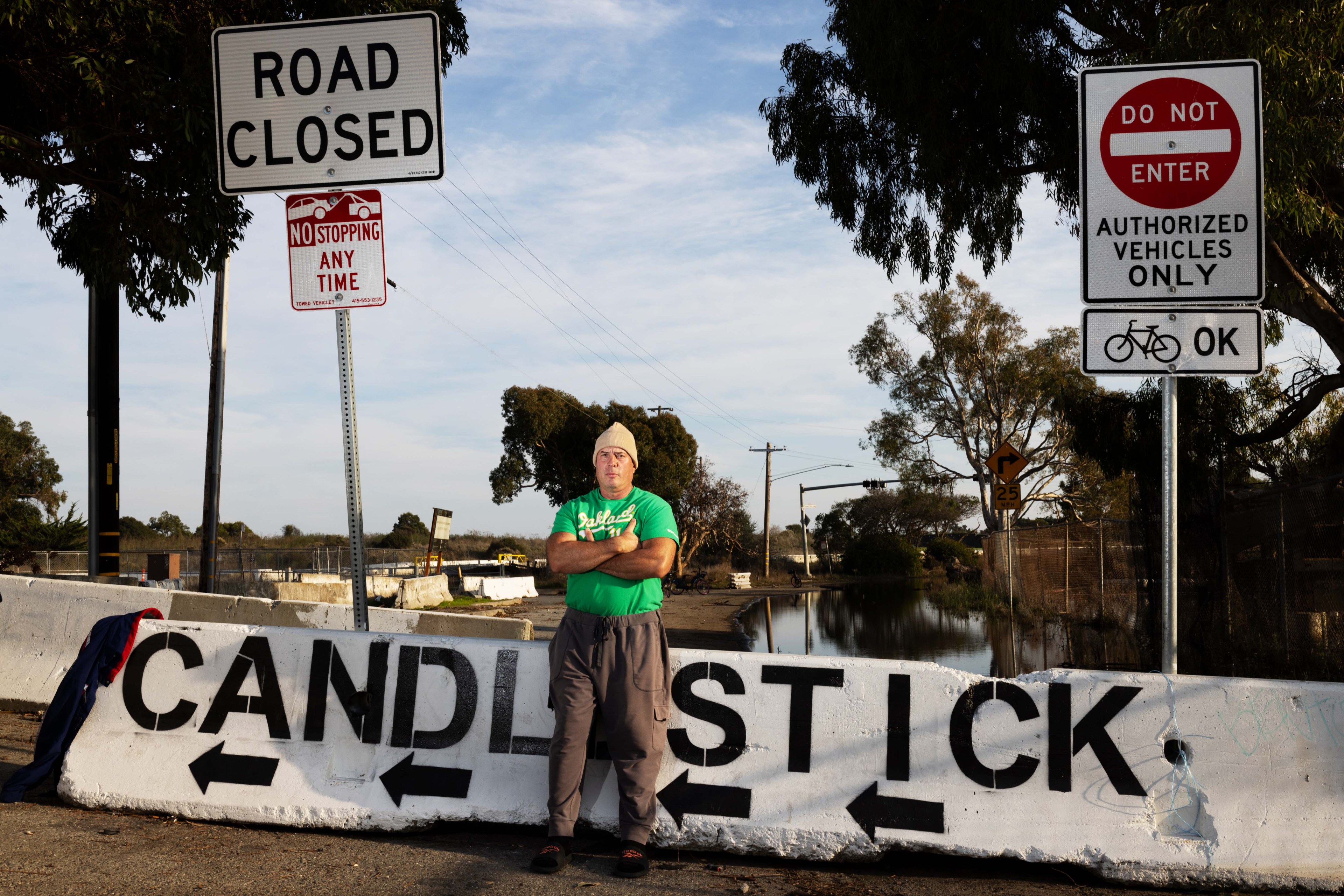 A person stands in front of a "Road Closed" and "Do Not Enter" signs, near a barrier labeled "CANDLESTICK." Trees and a flooded road are in the background.